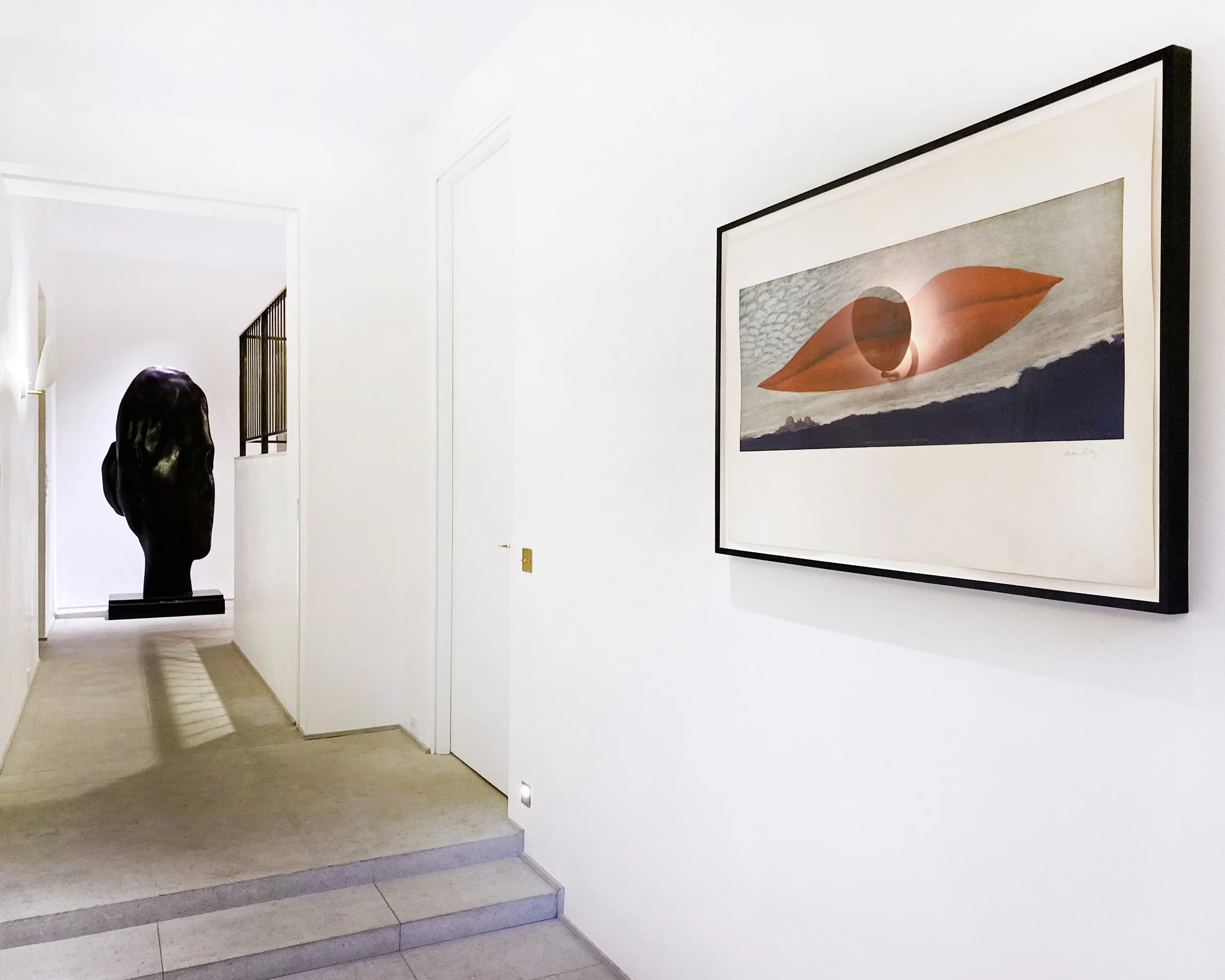 A hallway with white walls, a framed surreal artwork with a land and sky scene, and a black sculptural head sculpture at the end of the hallway.