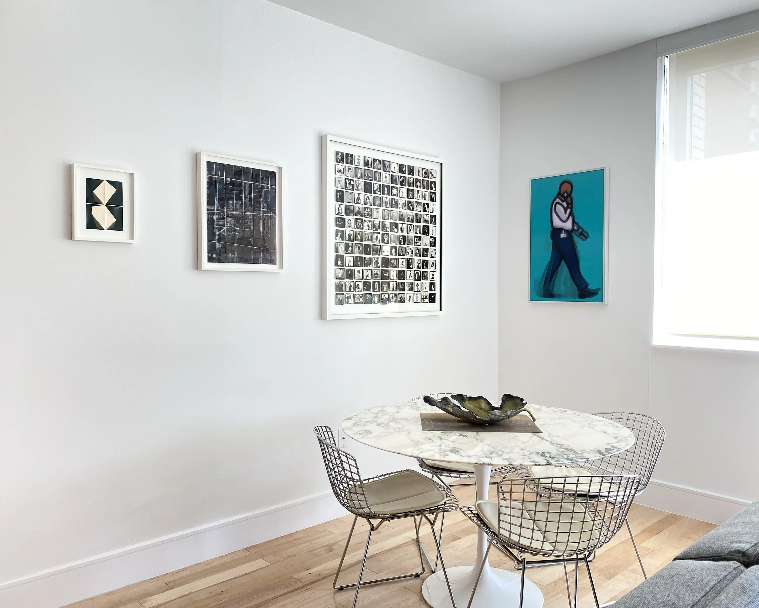 Modern dining area with a white marble round table, four wire chairs with cushions, framed artwork on white walls, and a large window letting in natural light.