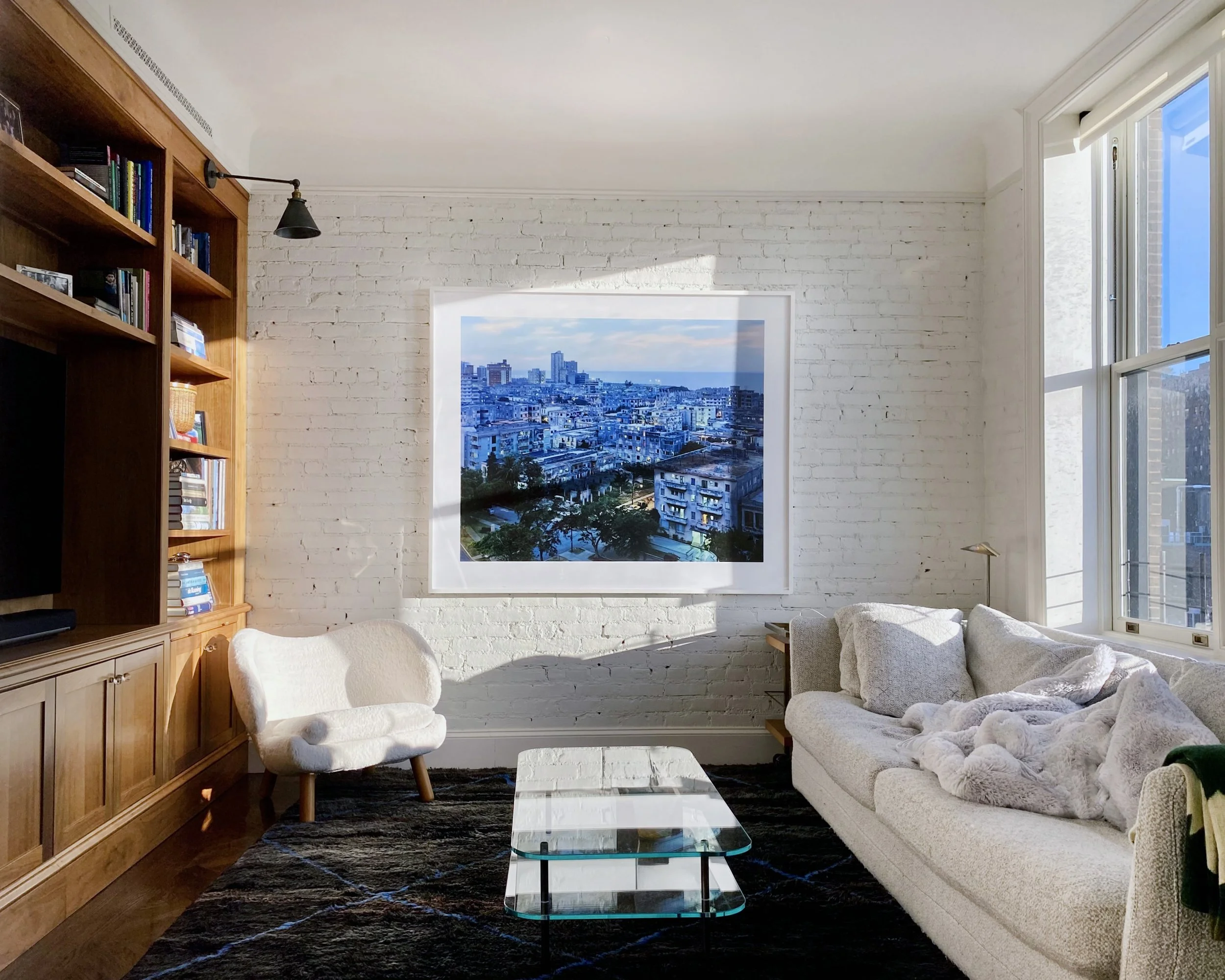 Living room with white brick wall, large window, artwork of a city skyline, gray sofa with blanket, white armchair, glass coffee table, dark rug, and wooden bookshelf.