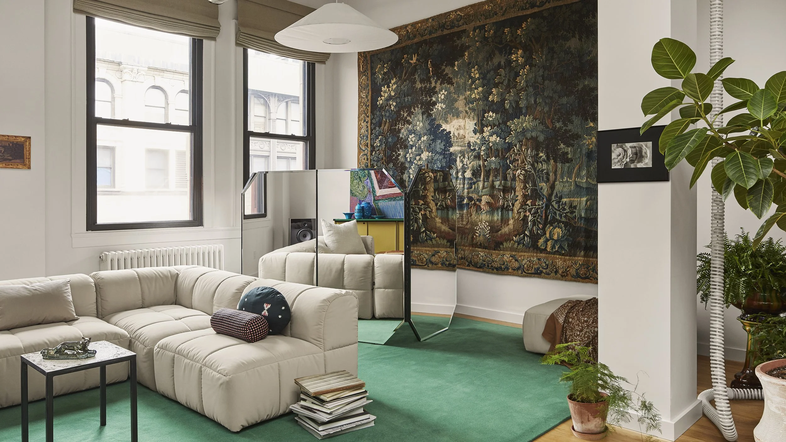 Living room with white sectional sofa, mirror, large tapestry wall hanging, potted plants, and windows.