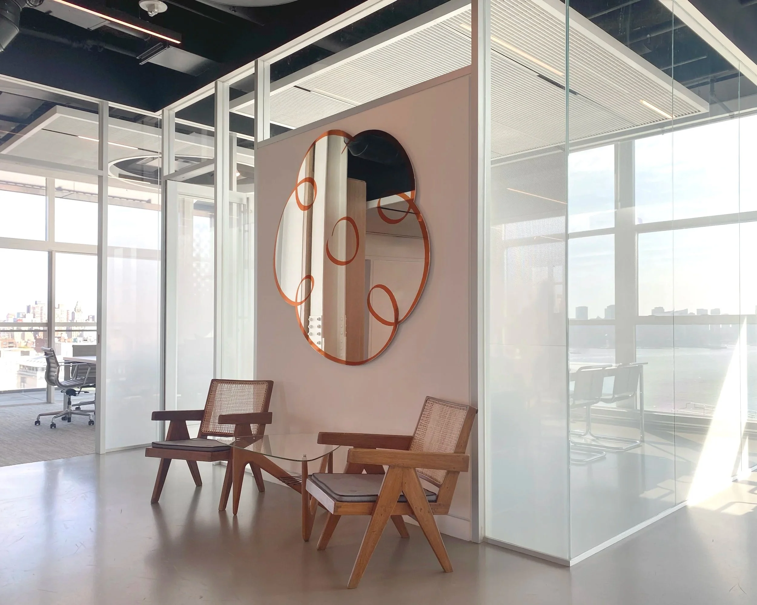Modern office interior with wooden chairs, glass table, and a wall mirror with orange circular design, large windows allowing natural light, and city skyline view.