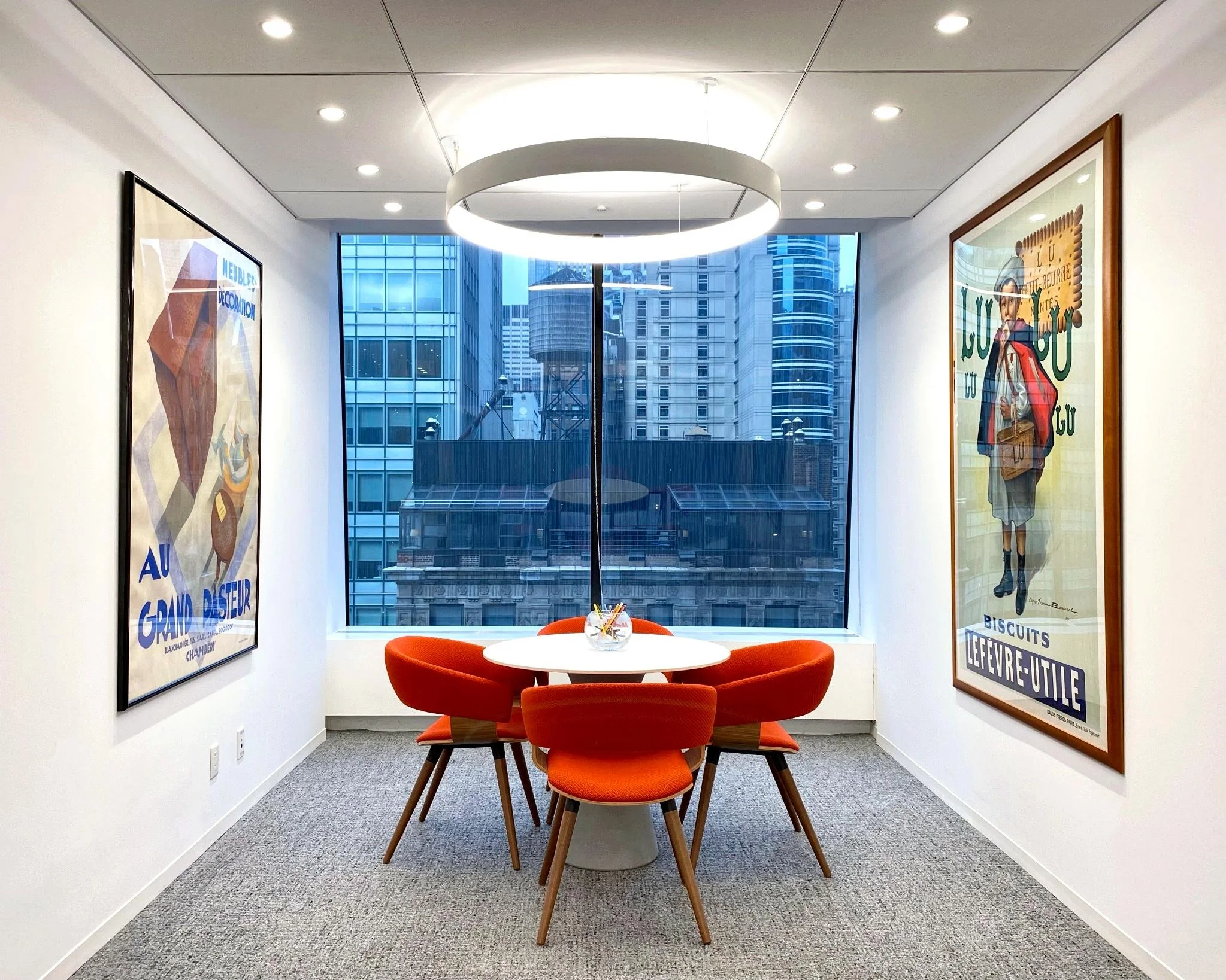 Modern office space with white walls and ceiling, decorated with framed vintage posters. Central round table with four orange upholstered chairs. Large window showing cityscape with tall glass buildings.