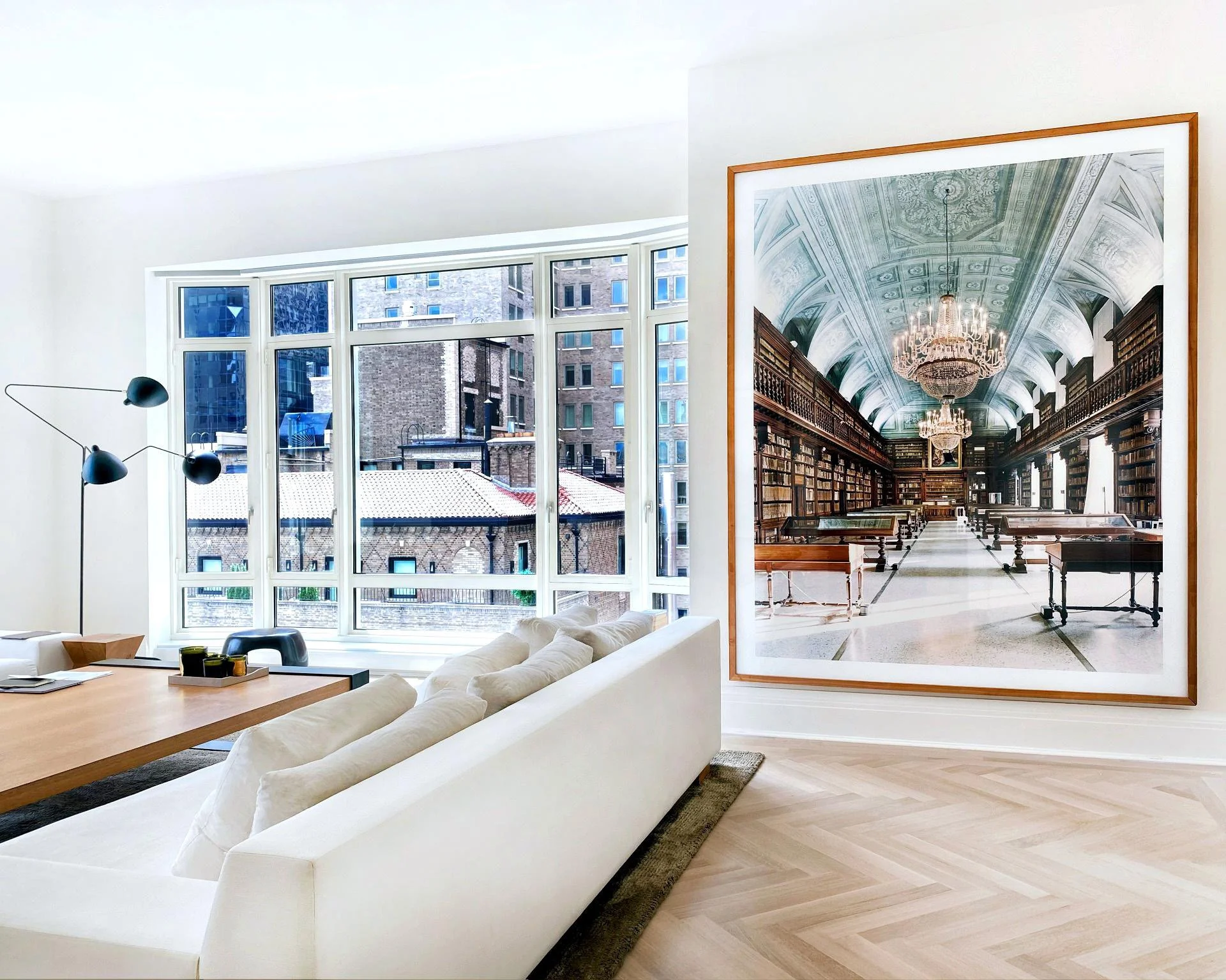 Modern living room with white sofa, large window showing city buildings, artwork of a grand library interior with chandeliers and bookshelves.