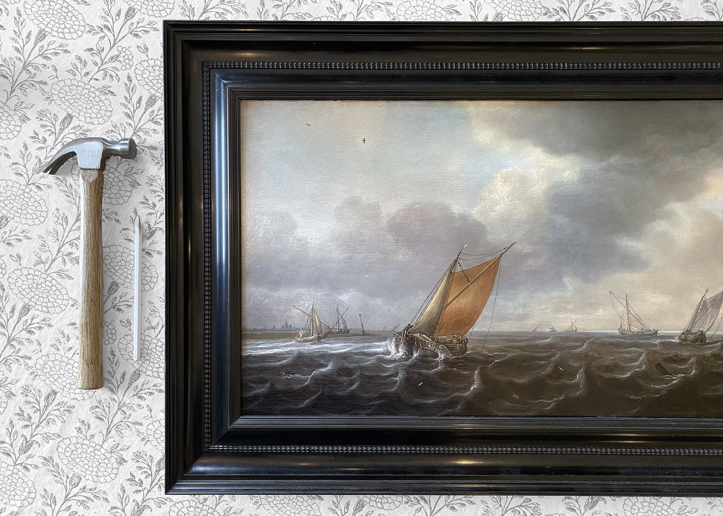 Close-up of a traditional oil painting of sailing ships on the ocean, framed in black, with a textured wallpaper background on the wall.