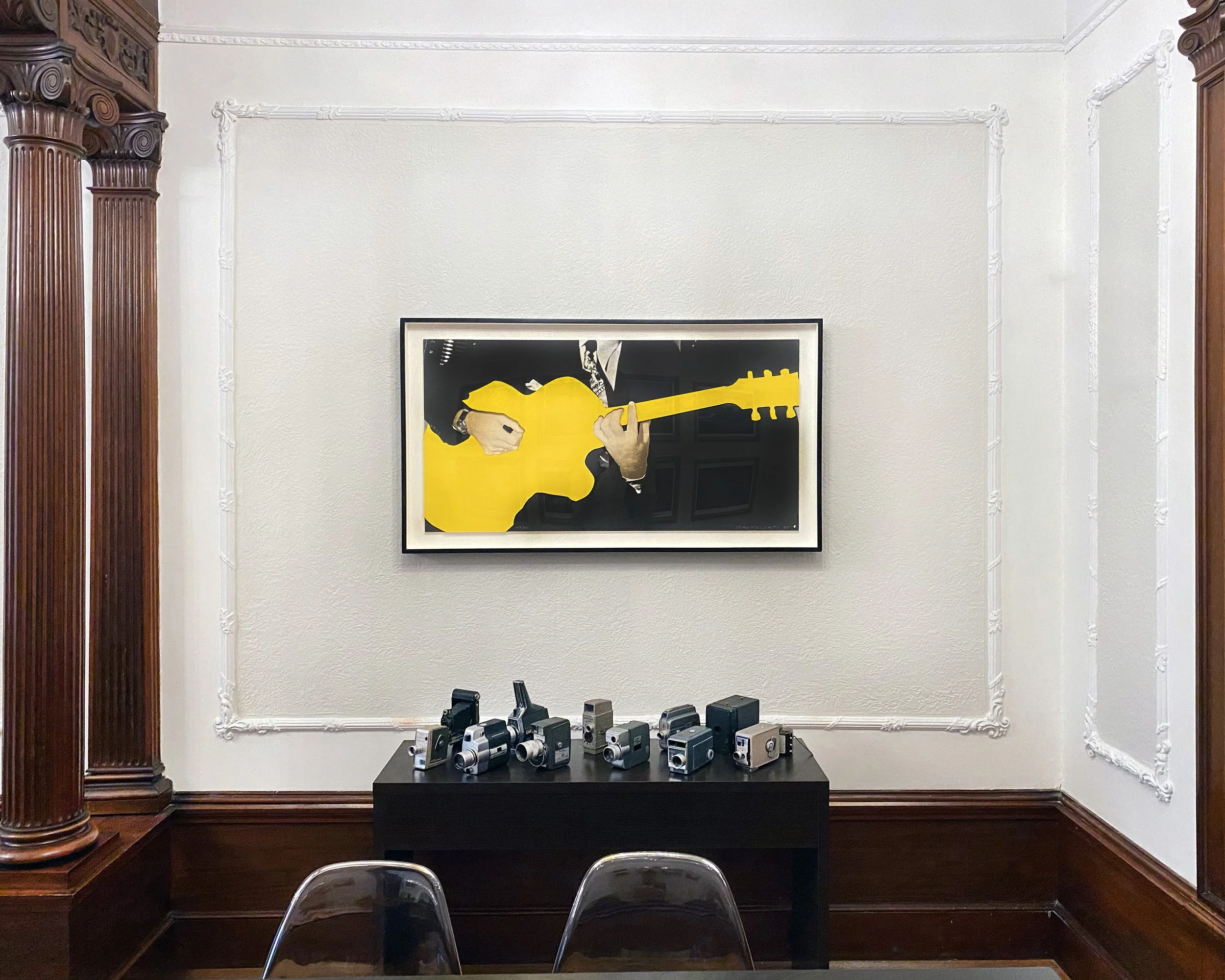 Gallery wall with a framed art piece of a person in a suit playing a yellow guitar. Below is a black table with vintage cameras on it and two clear plastic chairs in the foreground.