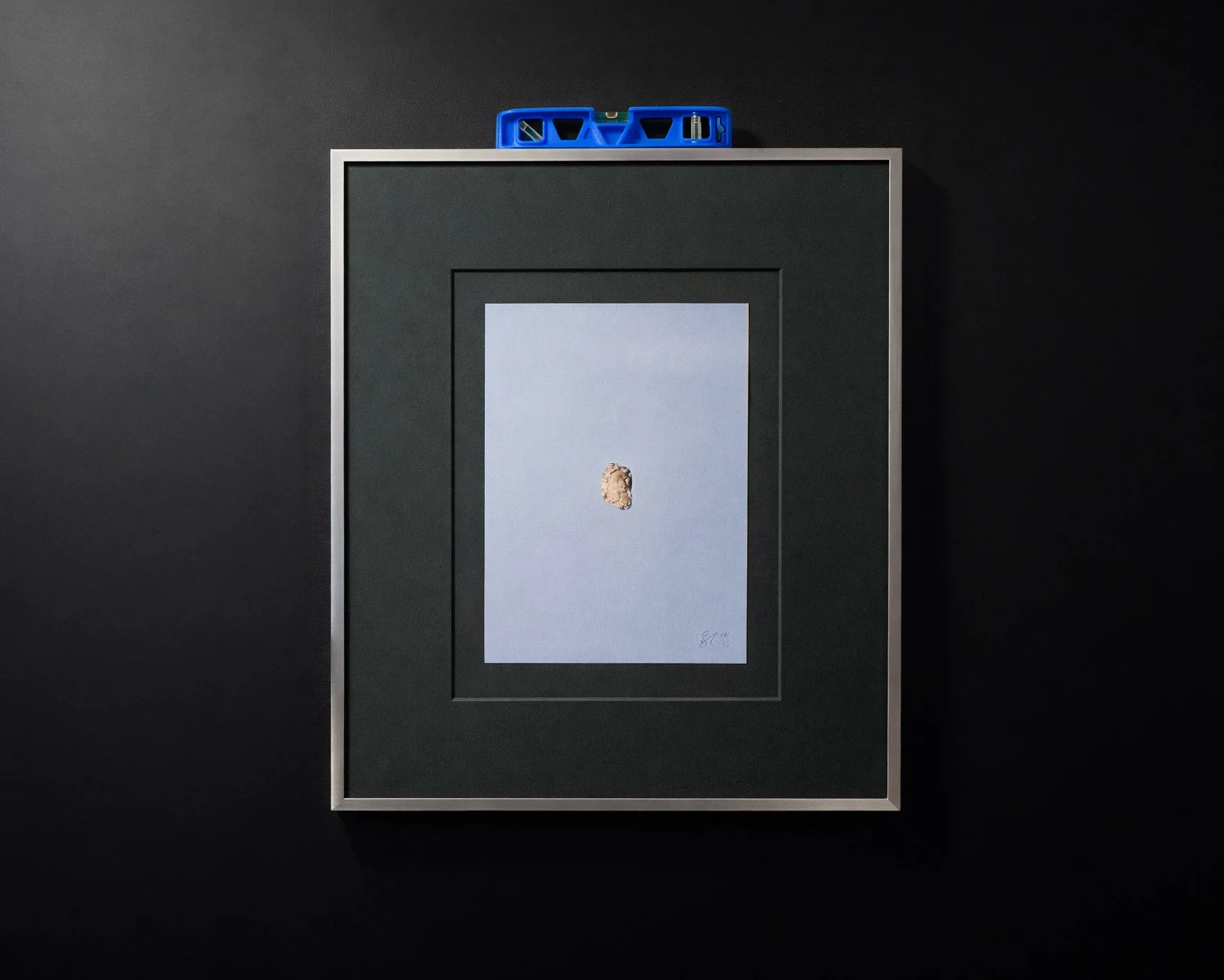 A framed specimen of a small rock or mineral against a light blue background, with a black and white border and a blue tool at the top of the frame.