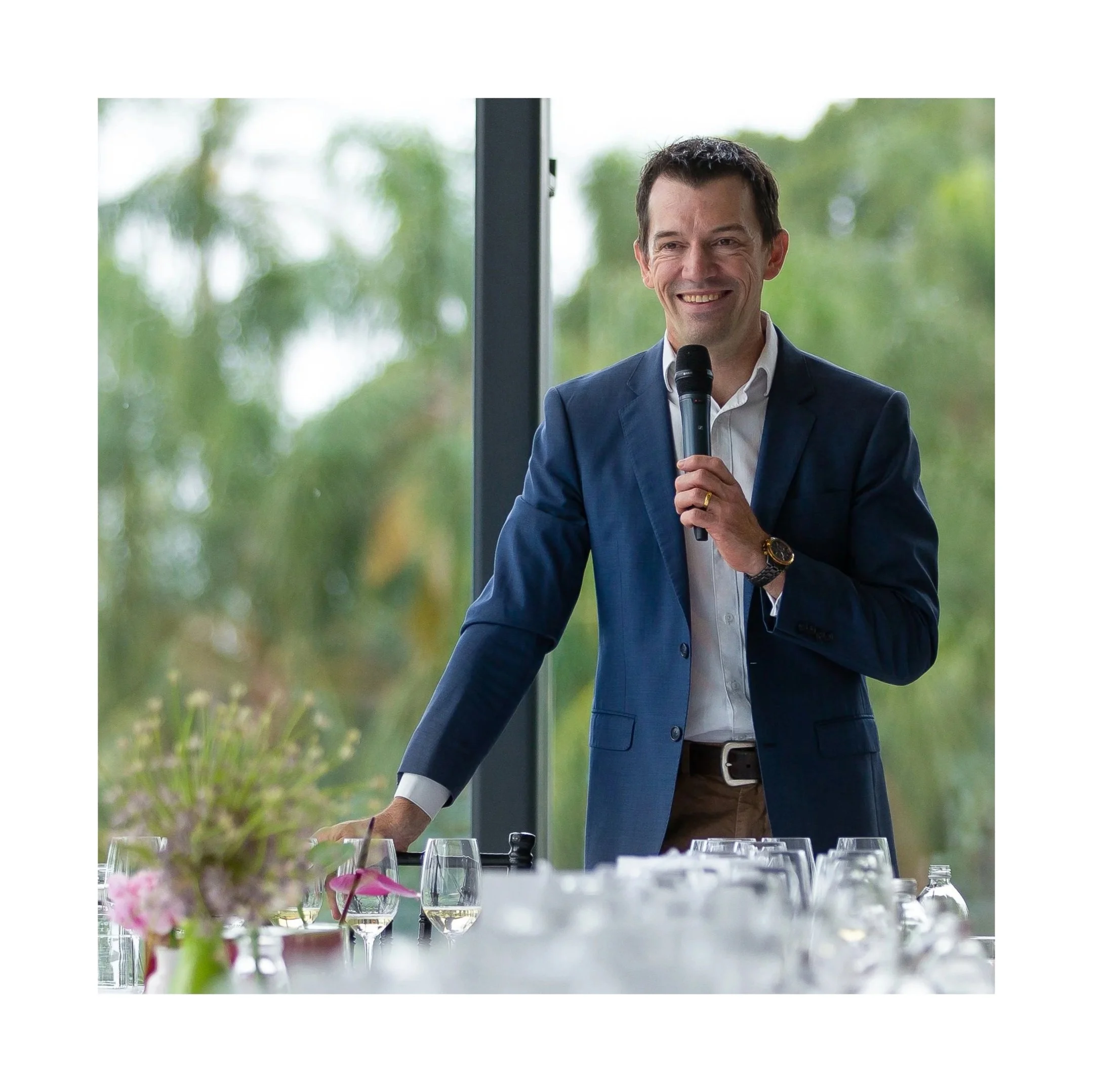 A man in a navy blue suit holding a microphone, standing at a table with wine glasses and flowers, smiling during a speech or presentation with greenery outside the window in the background.