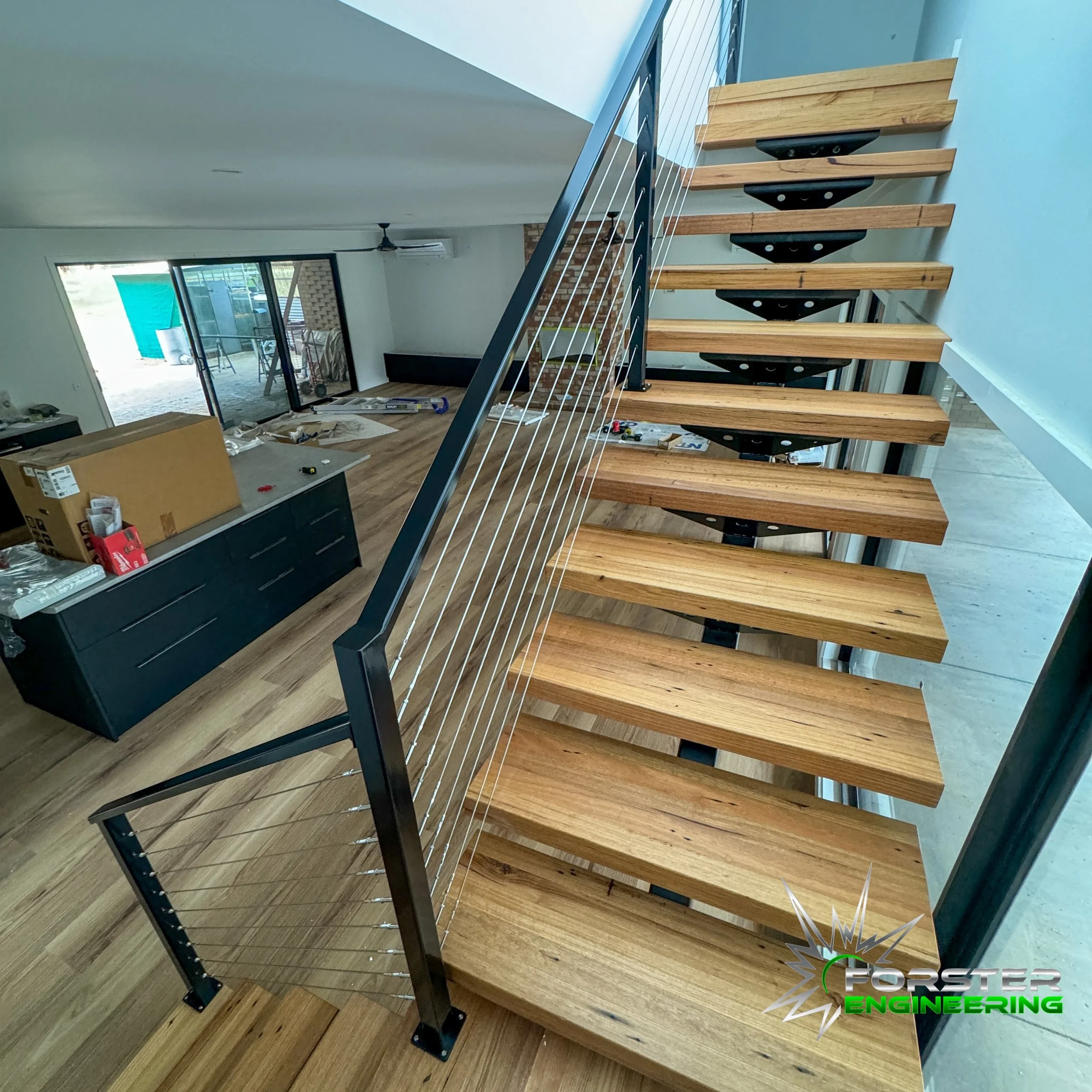 Interior view of a house under construction featuring a wooden staircase with metal supports, a black railing, and an open living area with large sliding glass doors, construction materials, and tools.