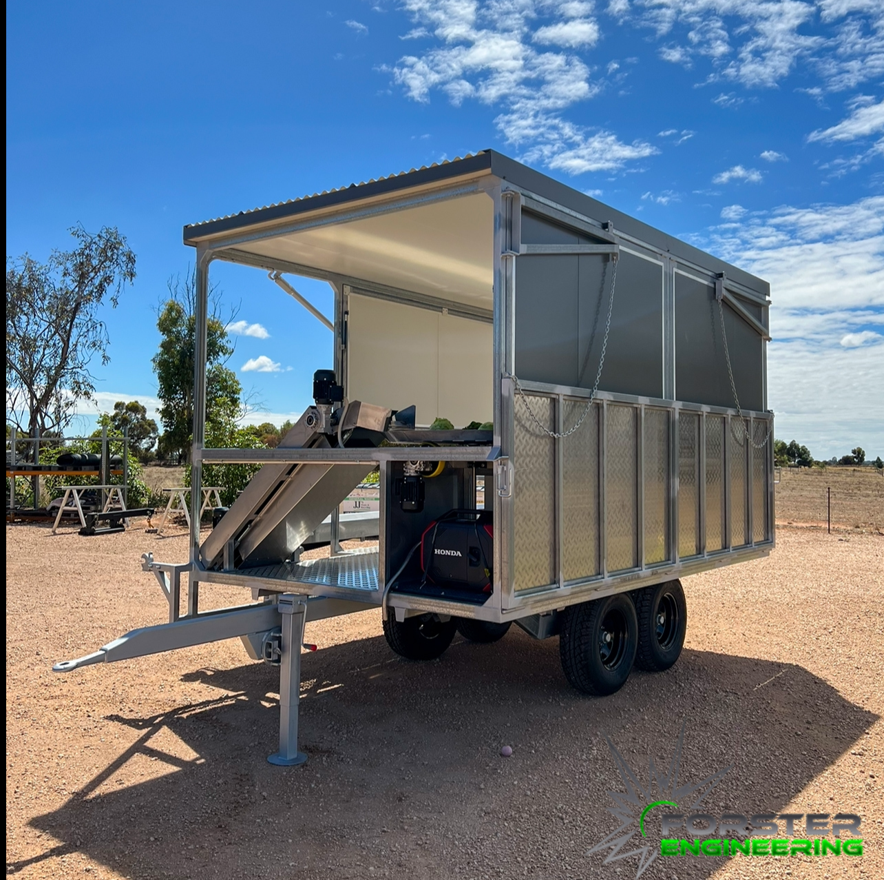 A modern mobile structure with an open front, minimal graphics, and a sloped roof, situated outdoors on a dirt surface with trees and a blue sky in the background, featuring the logo 'Gorster Engineering' at the bottom right.