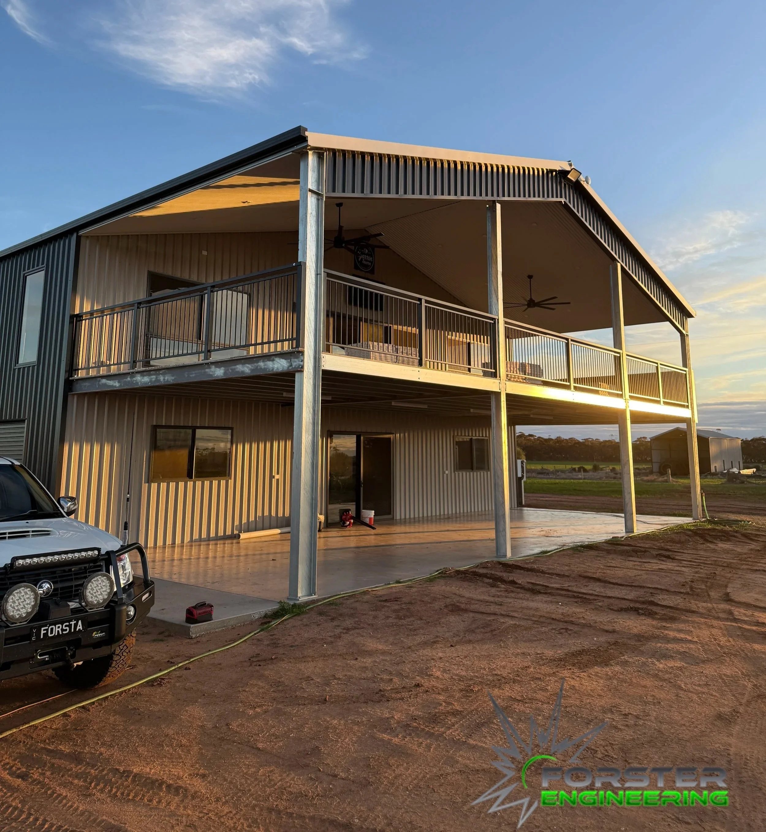 Two-story metal building with an open ground level and a balcony on the upper level, located in a rural area during sunset.