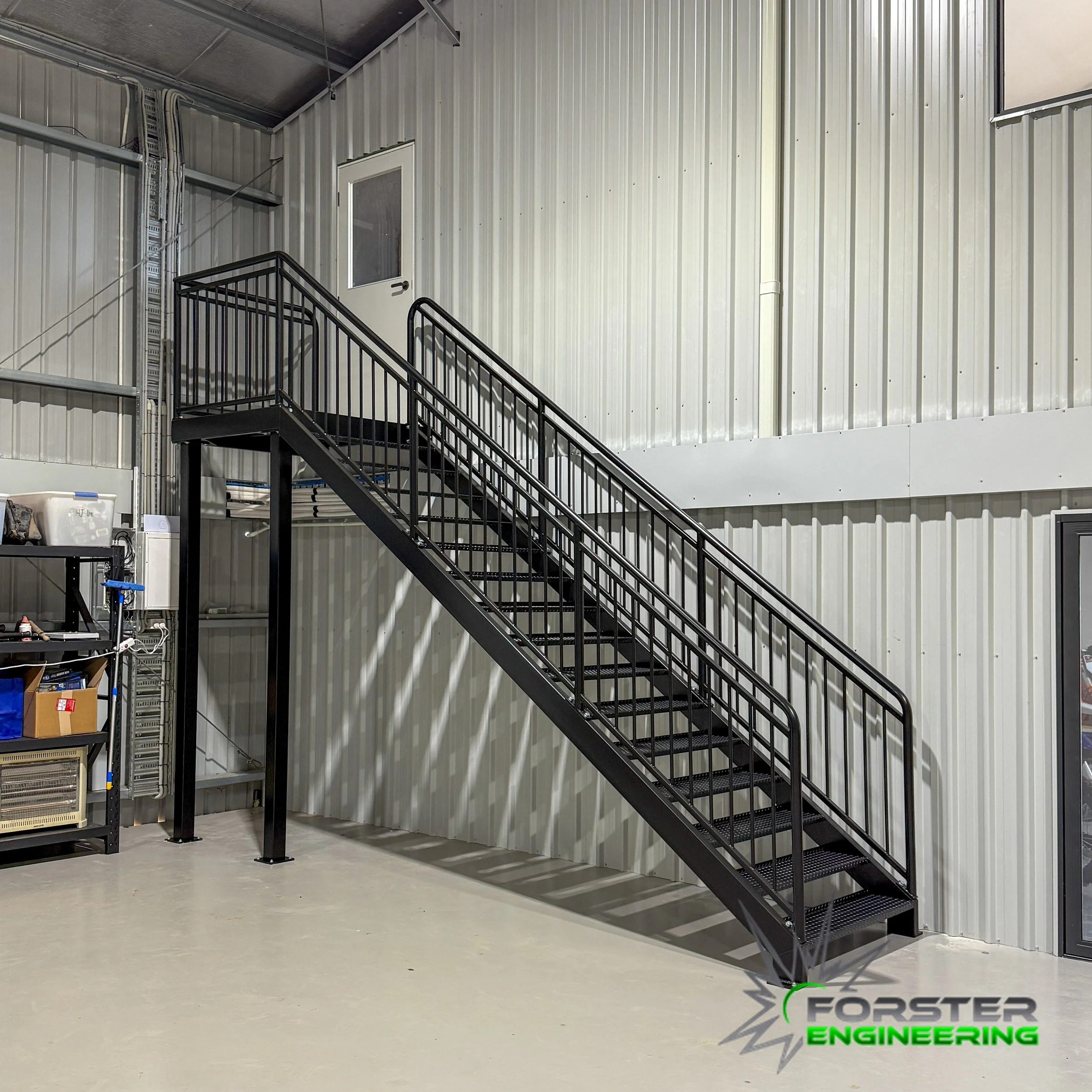 Interior of an industrial warehouse with a black metal staircase leading to a second-floor door. The walls are made of corrugated metal panels, and there are storage shelves on the left side. The floor is concrete. The Forster Engineering logo is in the bottom right corner.