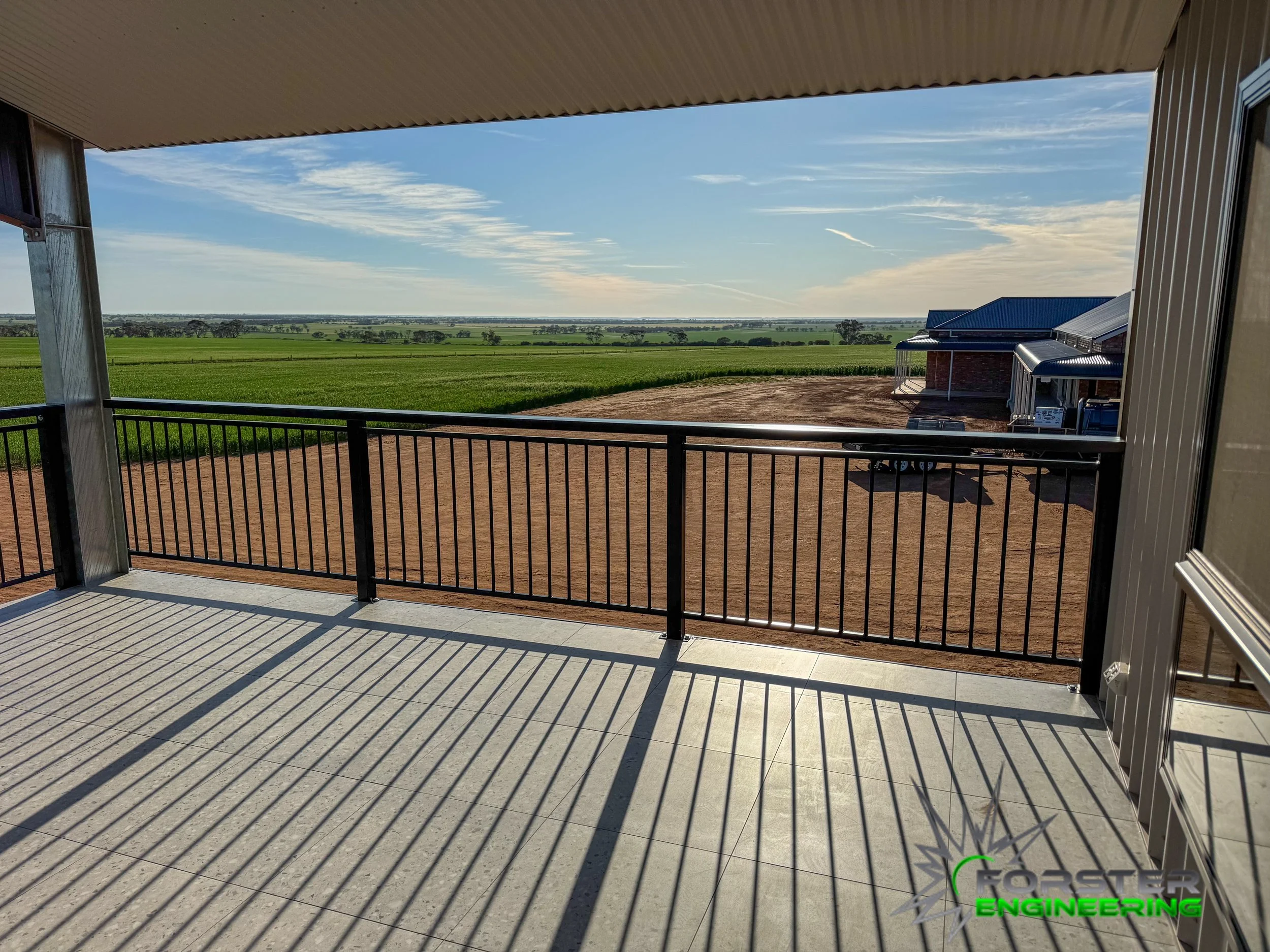 View from a balcony showing a green field, a dirt area, a blue sky with some clouds, and a building with a blue roof on the right side.