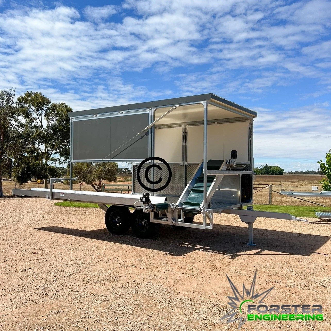 A Lettuce Harvesting Aid used by Farmers to Harvest their lettuce crops. Situated outdoors on a gravel surface with trees and open land in the background under a partly cloudy sky.