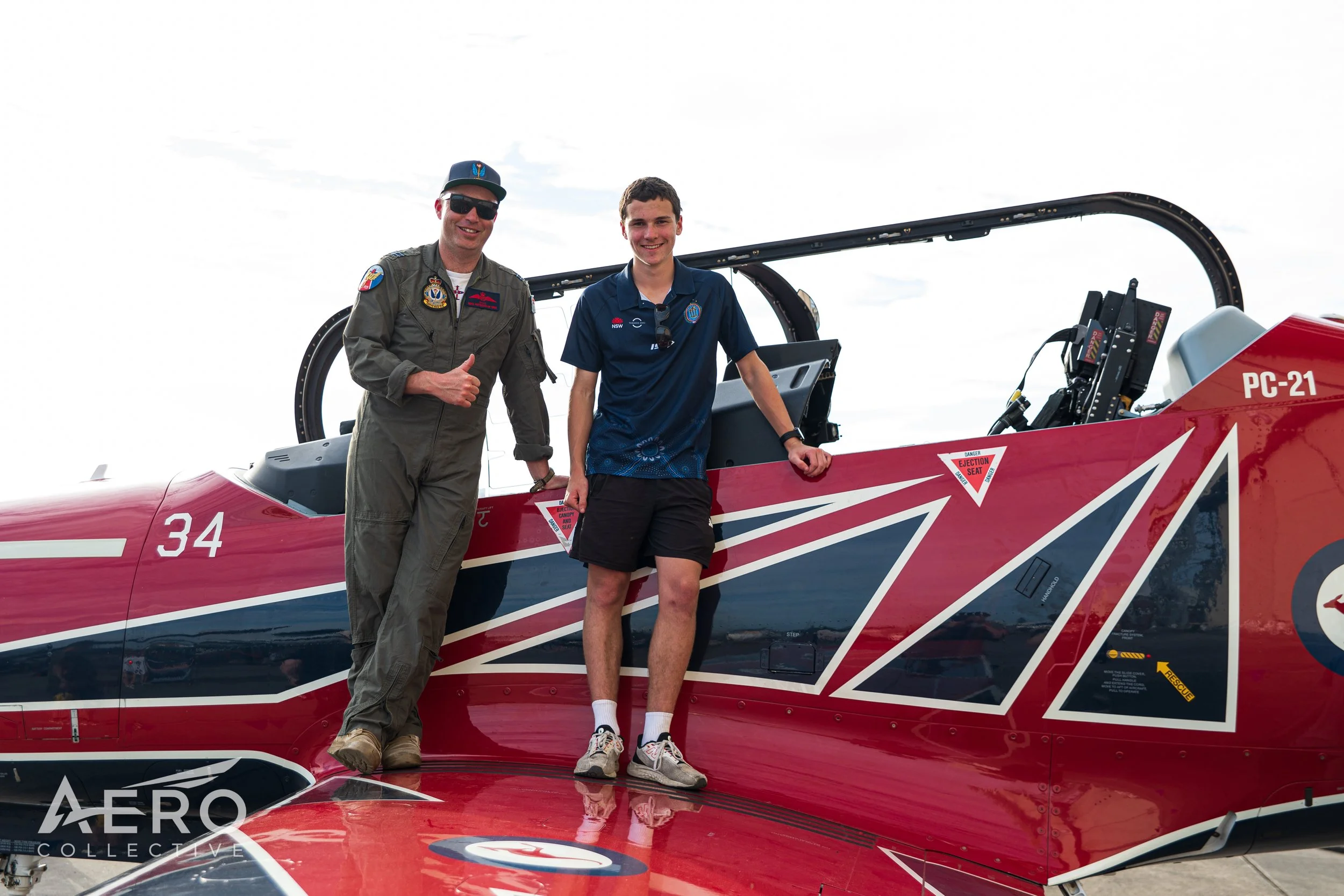 Two men standing beside a red fighter jet aircraft, smiling at the camera. One man is in a gray pilot jumpsuit, wearing sunglasses, and giving a thumbs-up. The other is in a blue polo shirt and black shorts, resting his arm on the jet.