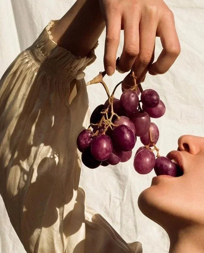 A person's hand holding a bunch of purple grapes close to their mouth, appearing to eat the grapes.
