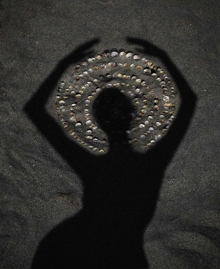 Person holding a circular object with concentric rings of small stones or beads, creating a spiral pattern, casting a shadow on a sandy surface.