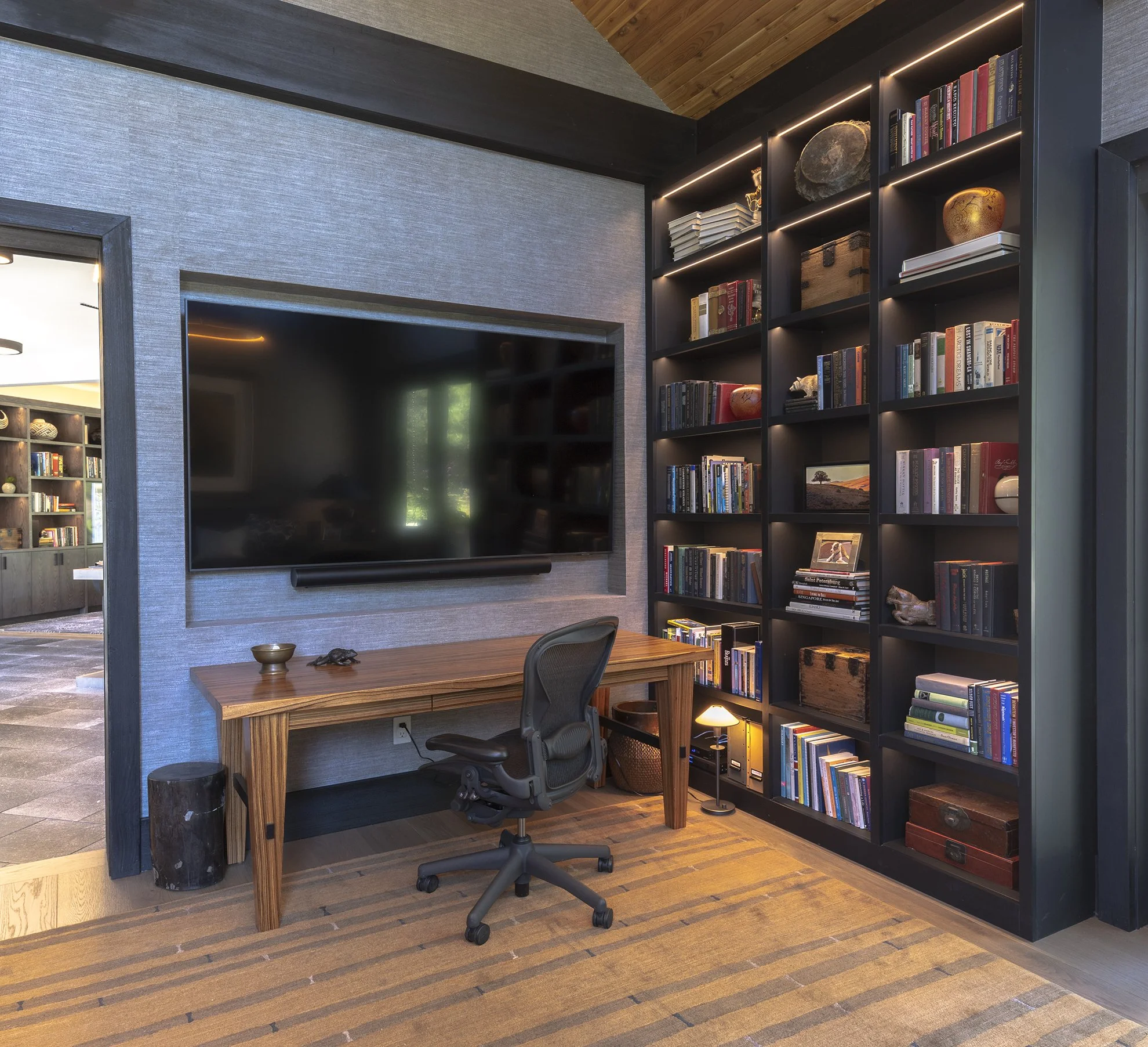 Interior view of Den with a recessed TV and library wall. The room features tongue and groove ceilings, fabric wall covering, and custom built shelves.