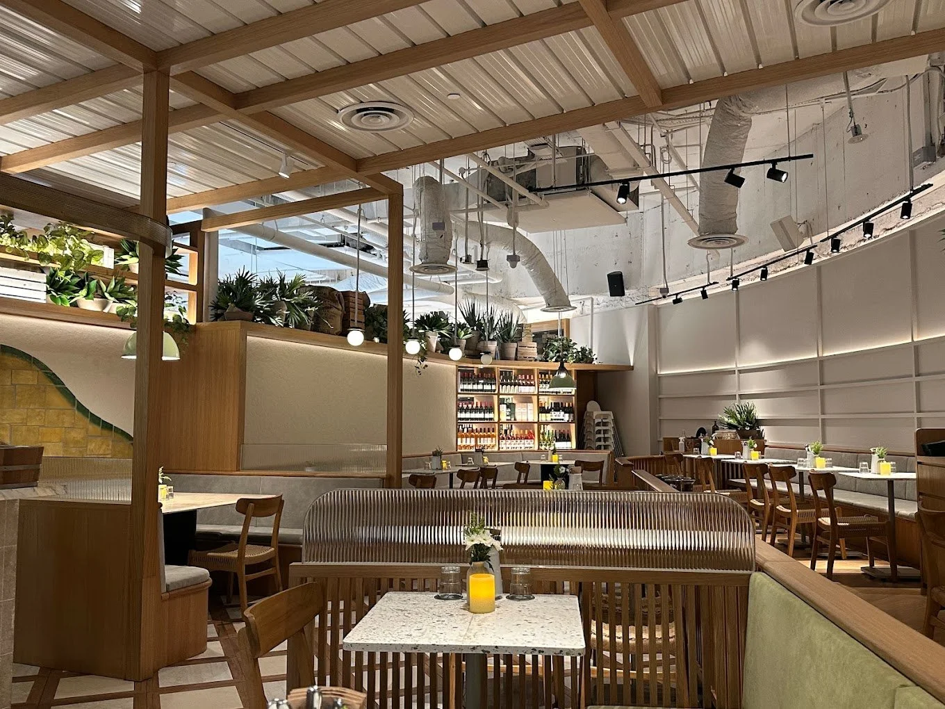 Interior of a modern restaurant or cafe with wooden furniture, plants, and white walls, featuring tables set with candles and small vases with flowers.