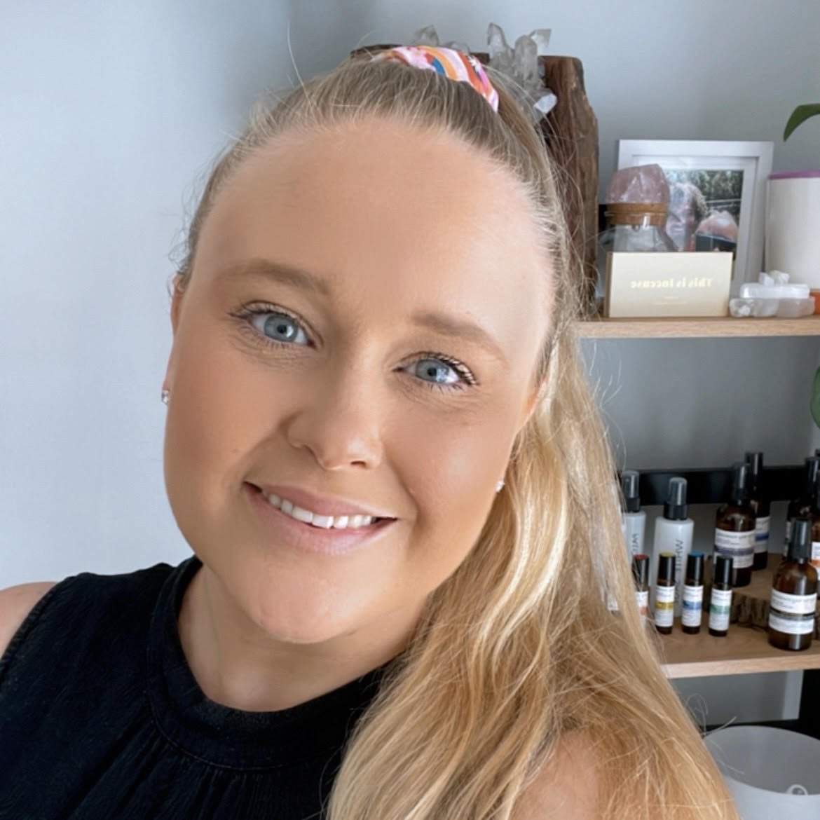 Close-up of a smiling woman with blue eyes, long blonde hair, and a colorful hair tie, indoors with shelves containing skincare products in the background.