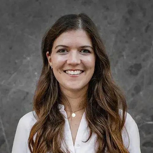 A woman with long, wavy brown hair and a white blouse, smiling in front of a gray textured background.