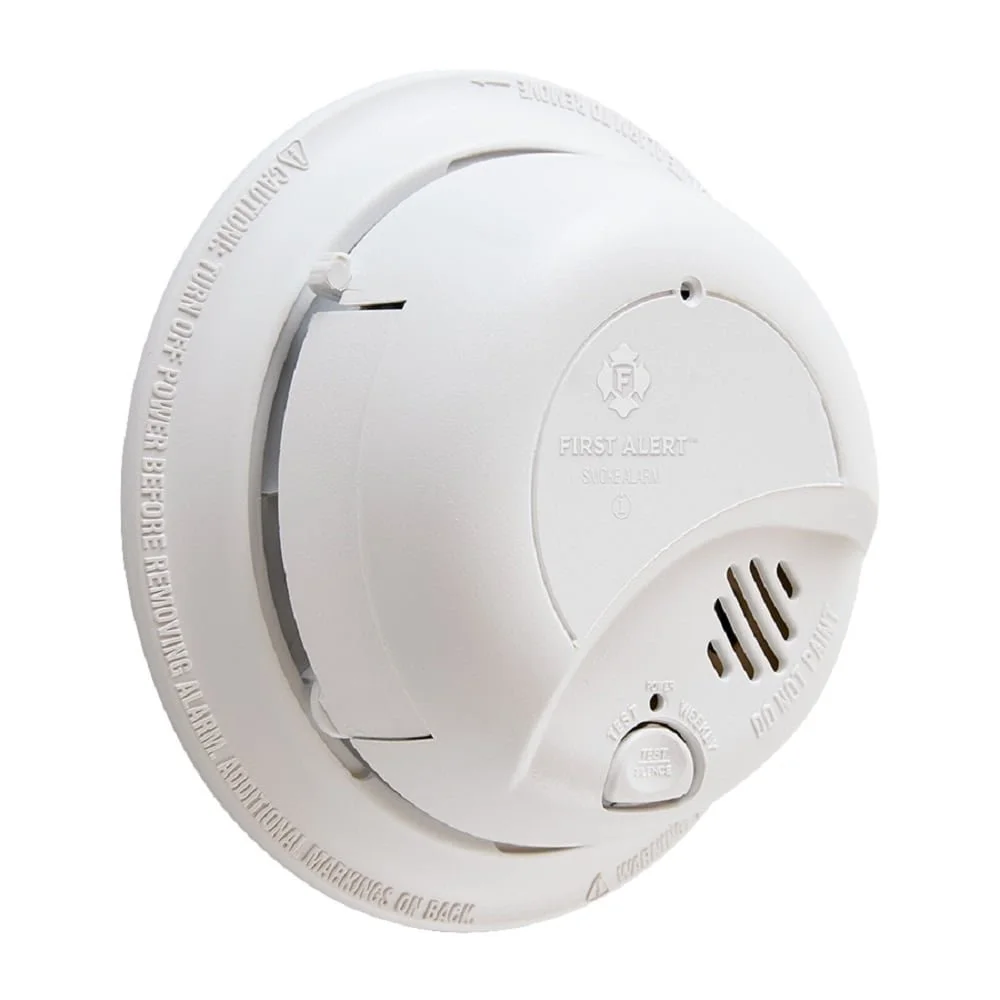 Smoke detector with First Alert branding, featuring a test button and vent slots, mounted on a ceiling.