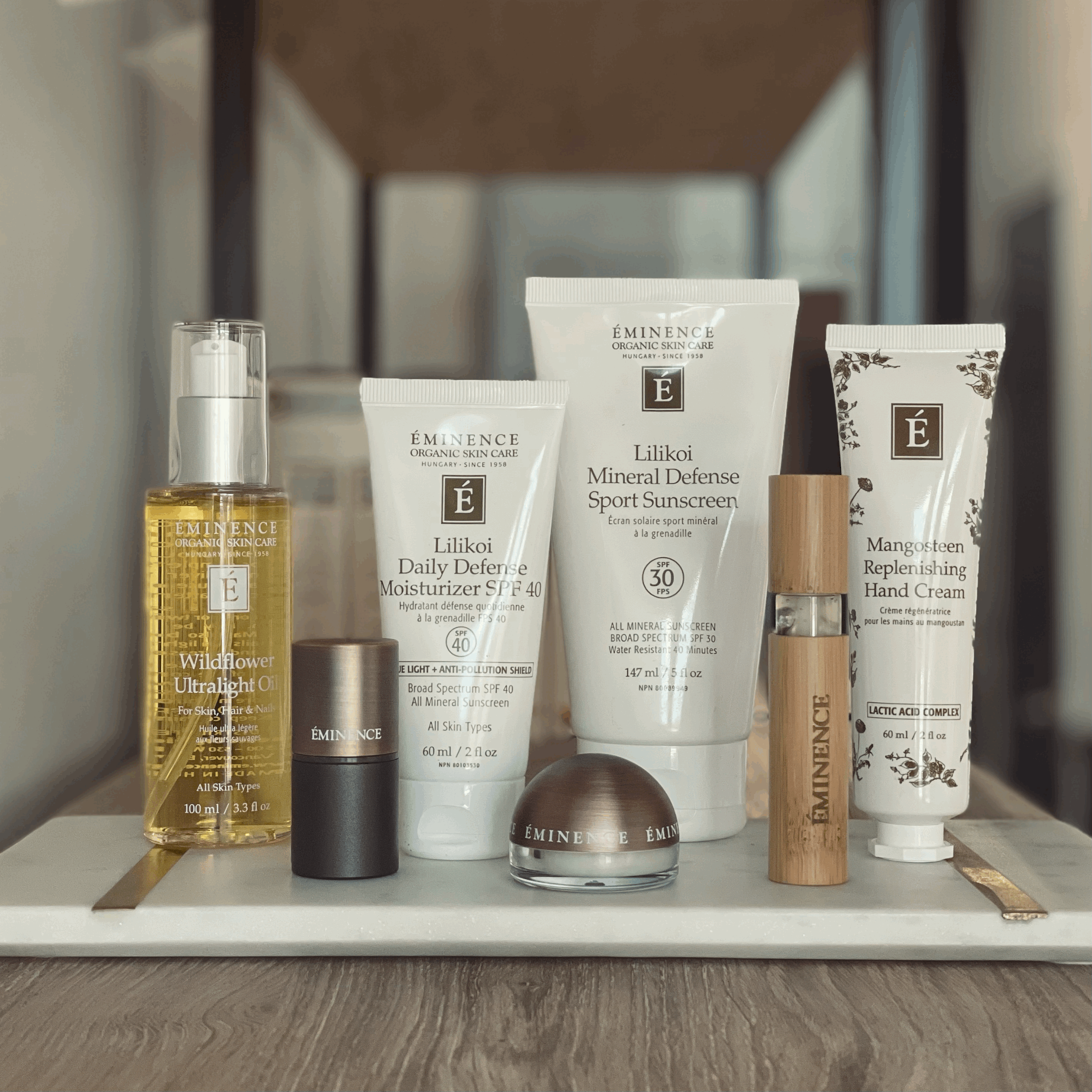 A collection of Eminence Organic Skin Care products on a tray, including Wildflower Ultralight Oil, Lilikoi Daily Defense Moisturizer SPF 40, Lilikoi Mineral Defense Sport Sunscreen, Mangosteen Replenishing Hand Cream, and a wooden-capped serum bottle, displayed indoors.