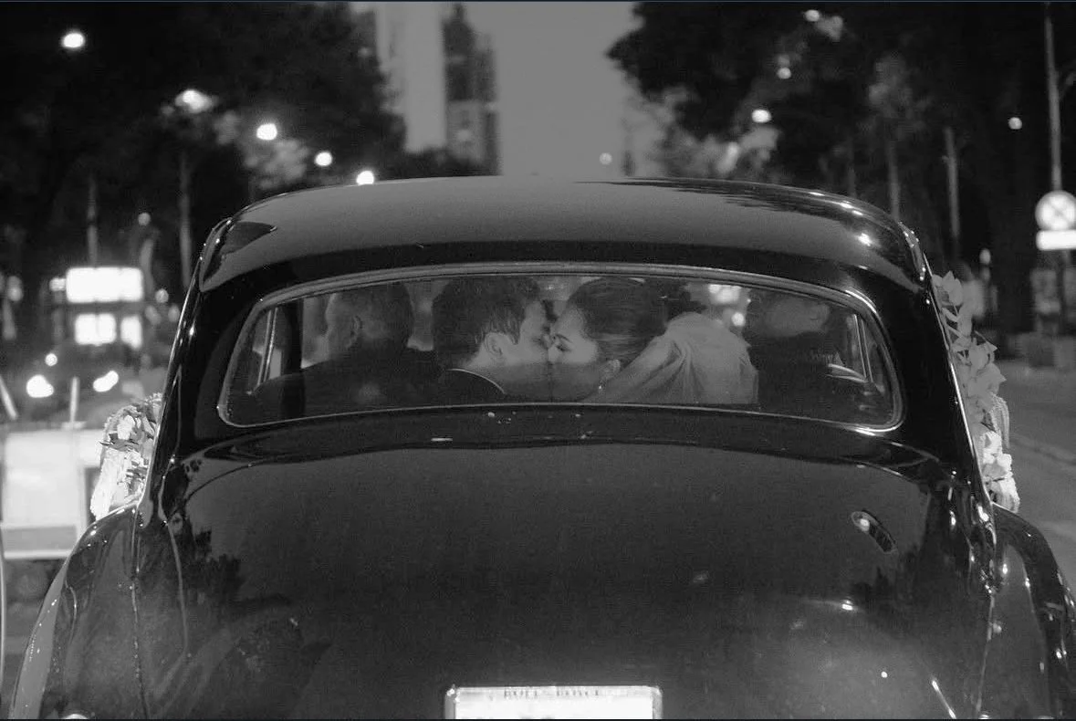 Rear view of a vintage car with a couple kissing inside; nighttime city scene with blurred lights in the background.