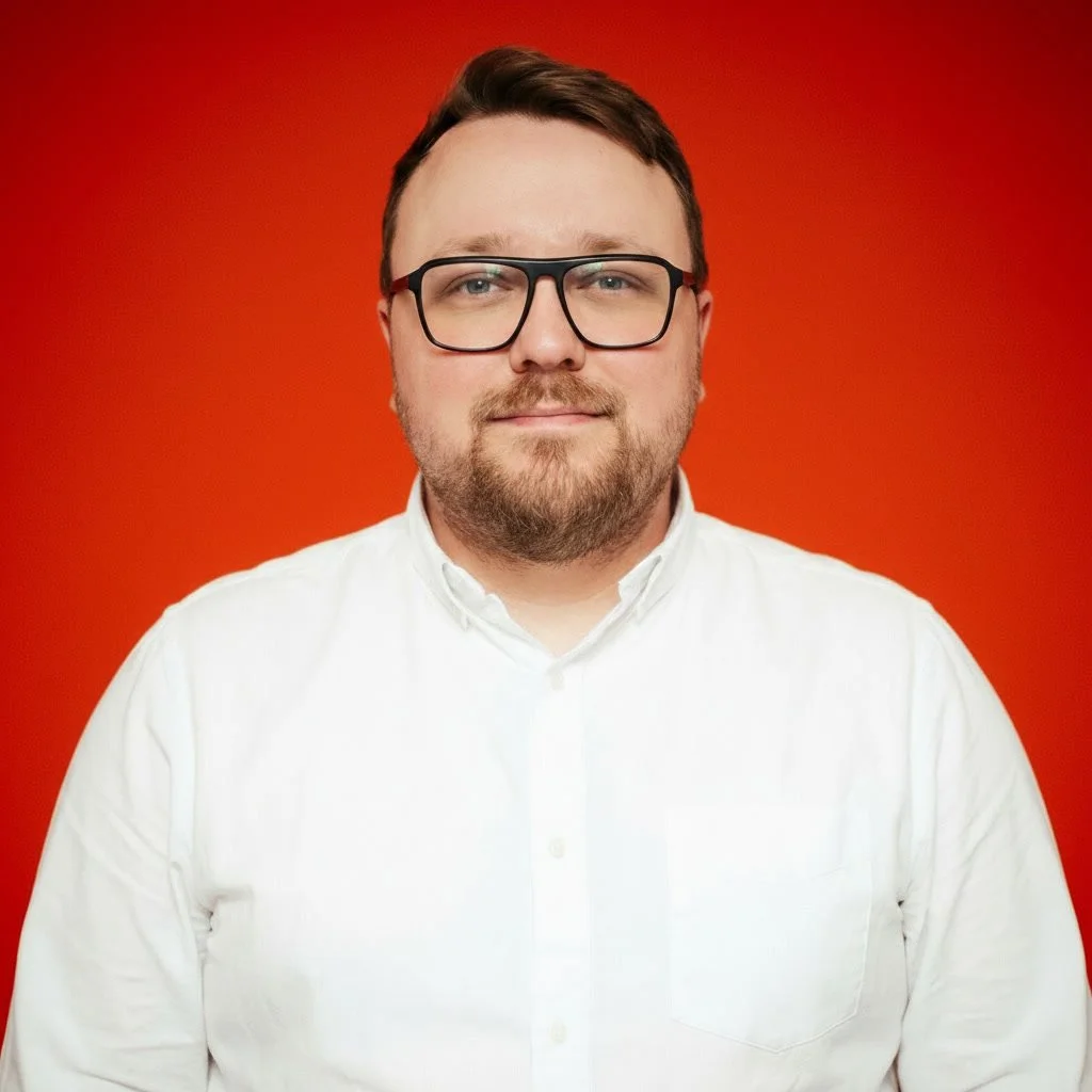Ein Mann mit Brille, weißen Hemd und lockigem Haar vor orangefarbenem Hintergrund, lächelnd mit verschränkten Armen.
