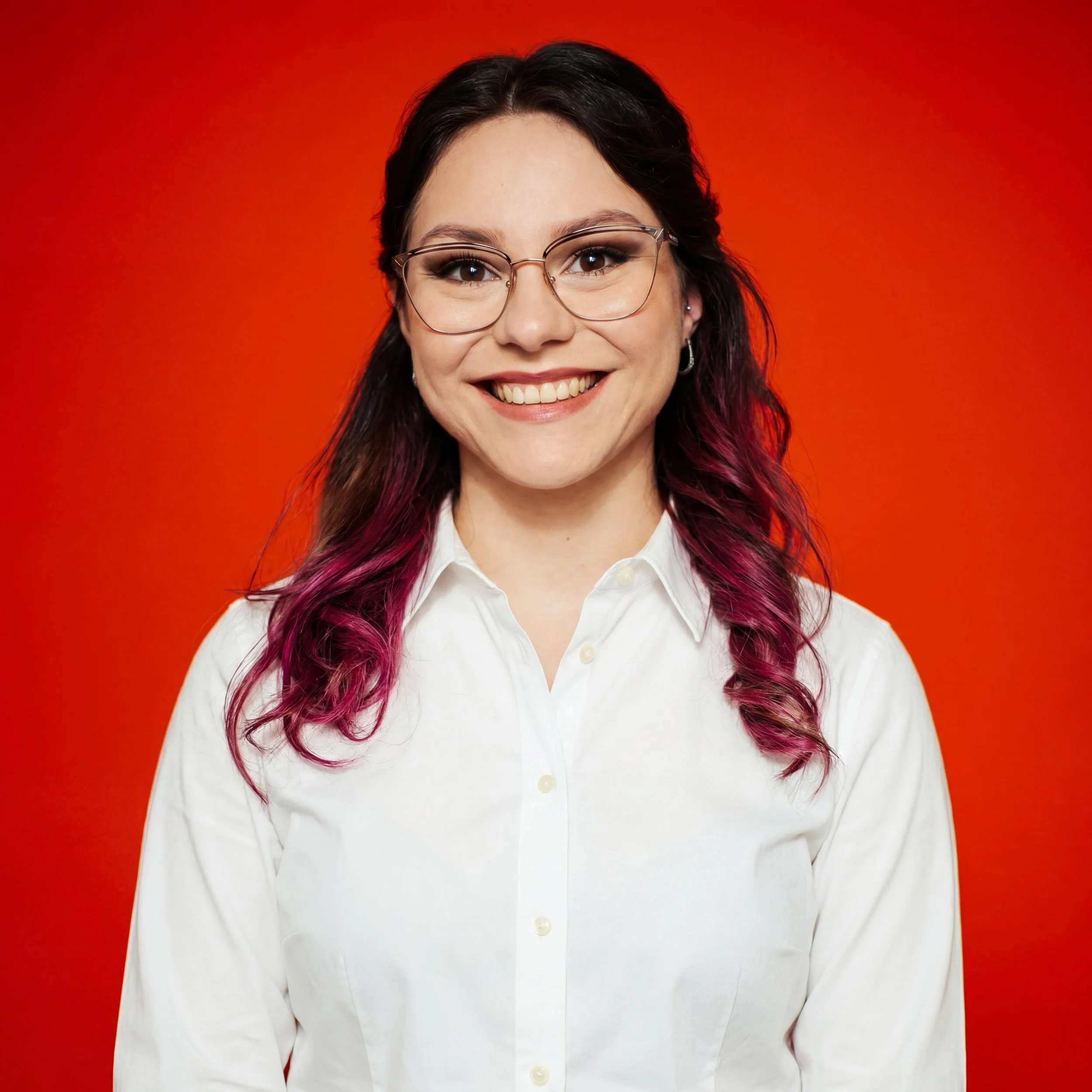 Eine Frau mit dunklen, lockigen Haaren, trägt eine Brille mit dünnem Rahmen und ein weißes Hemd, steht vor einem orangefarbenen Hintergrund.