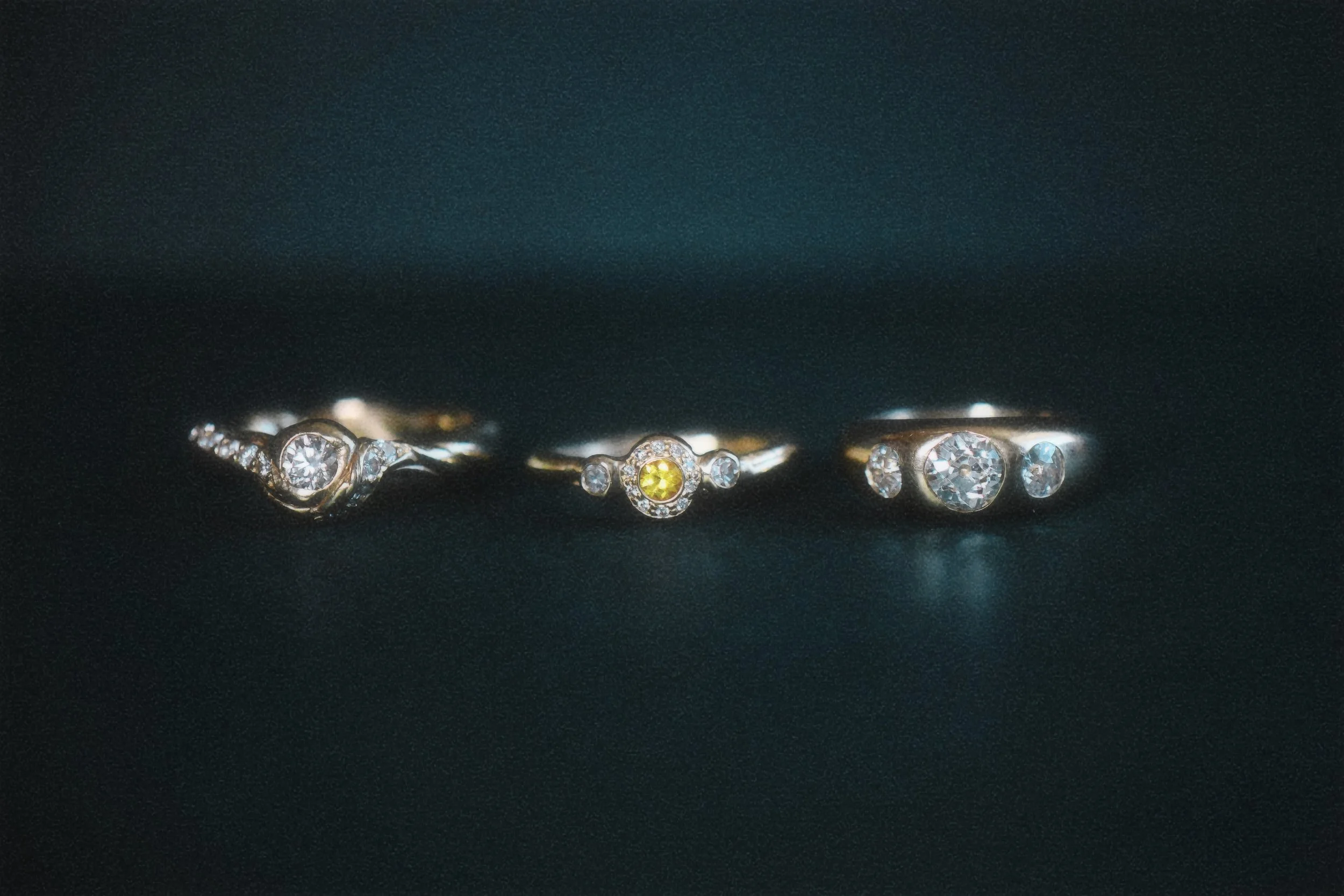 Three rings laid on a dark surface, each featuring a prominent central diamond or gemstone, with intricate designs and additional small stones around the main stones.
