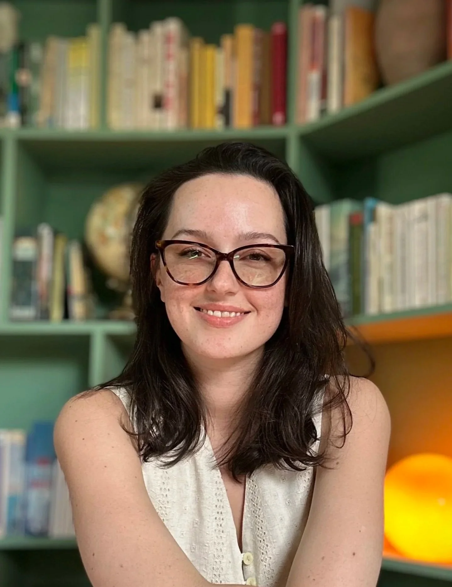 A young woman with light skin, brown hair, and freckles, smiling in a bright indoor setting with large windows and blurred background decor.