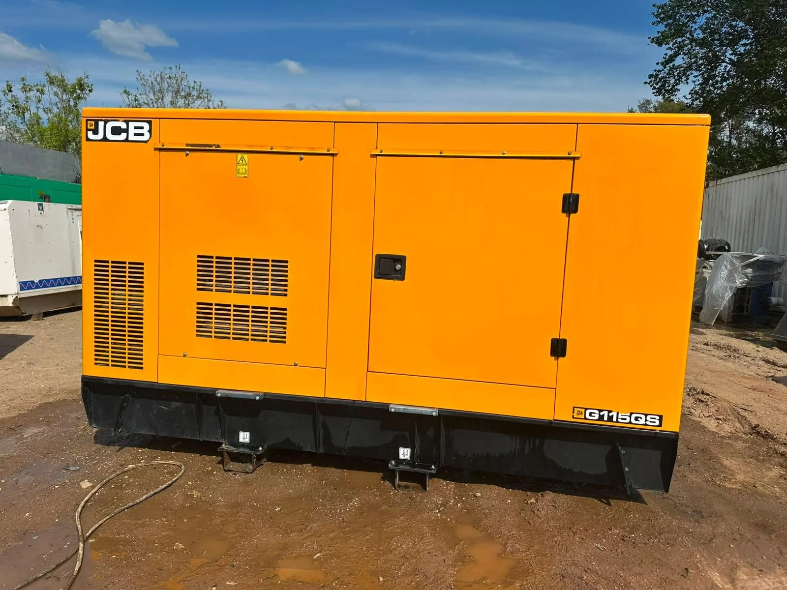 Yellow portable generator with JCB and G115QS labels, situated outdoors on muddy ground, with a partly cloudy blue sky and trees in the background.
