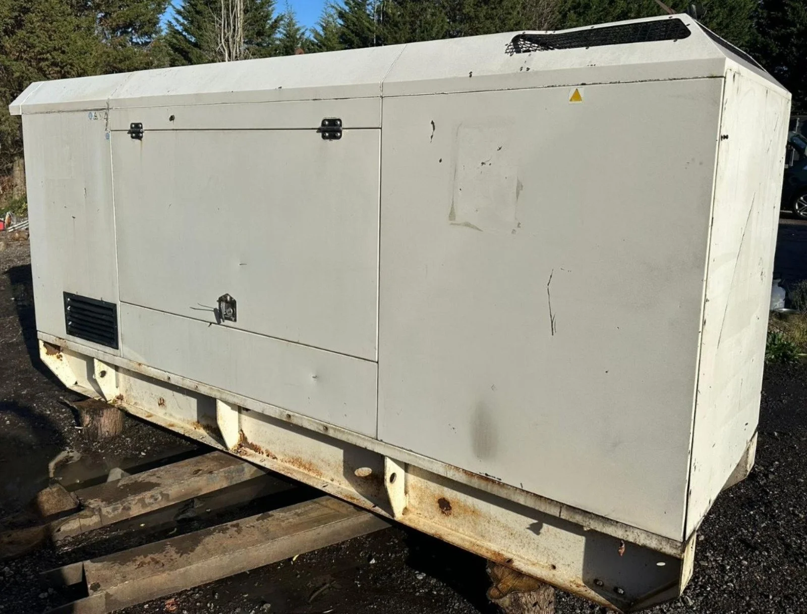 An outdoor industrial generator on a rusty metal frame, with a white metal casing, located on a dirt surface.