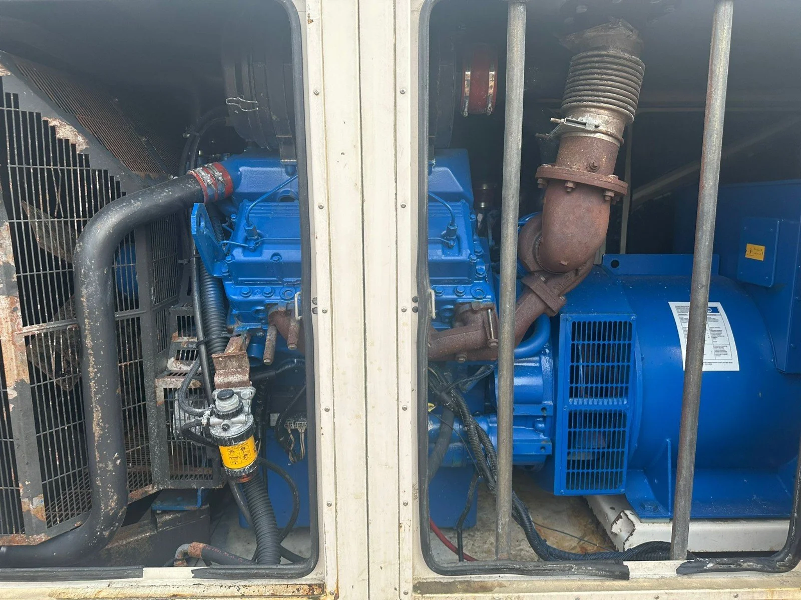 An industrial generator with a visible blue engine, rusted exhaust pipe, and various hoses and parts enclosed in a metal frame.