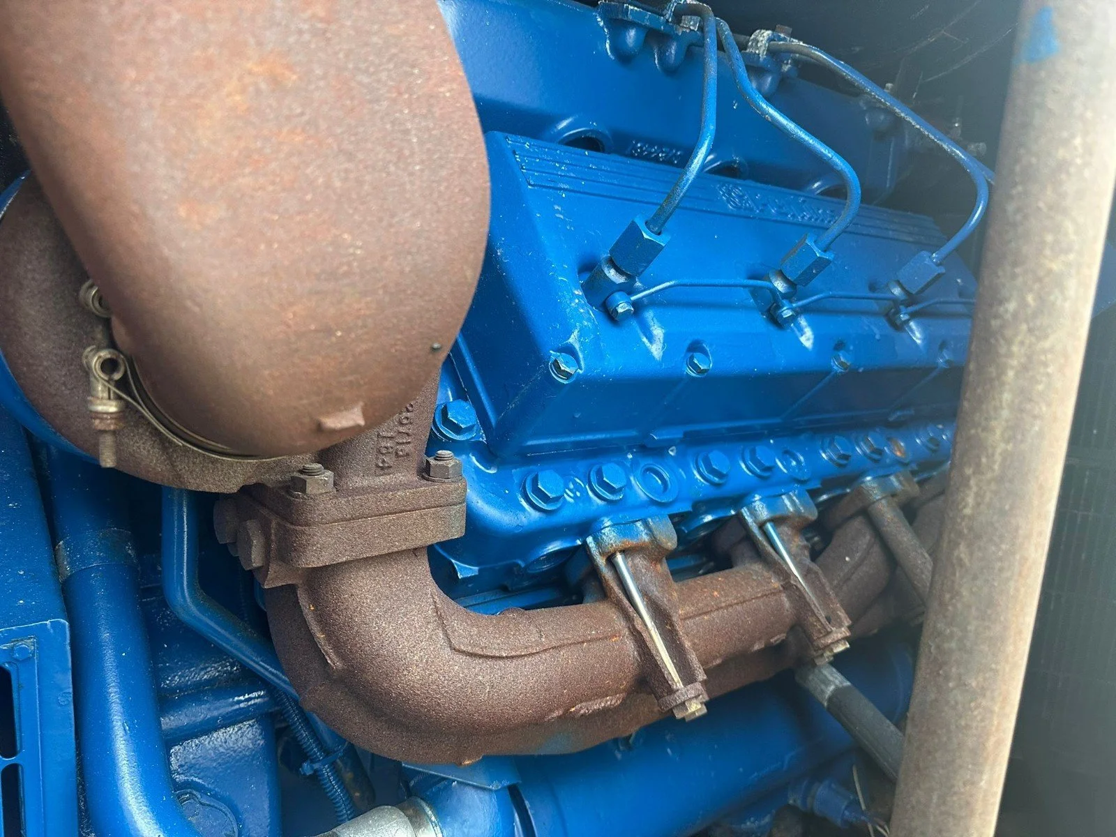Close-up of a bright blue industrial machine engine with metal pipes and components, including a rusty exhaust manifold on the left side.