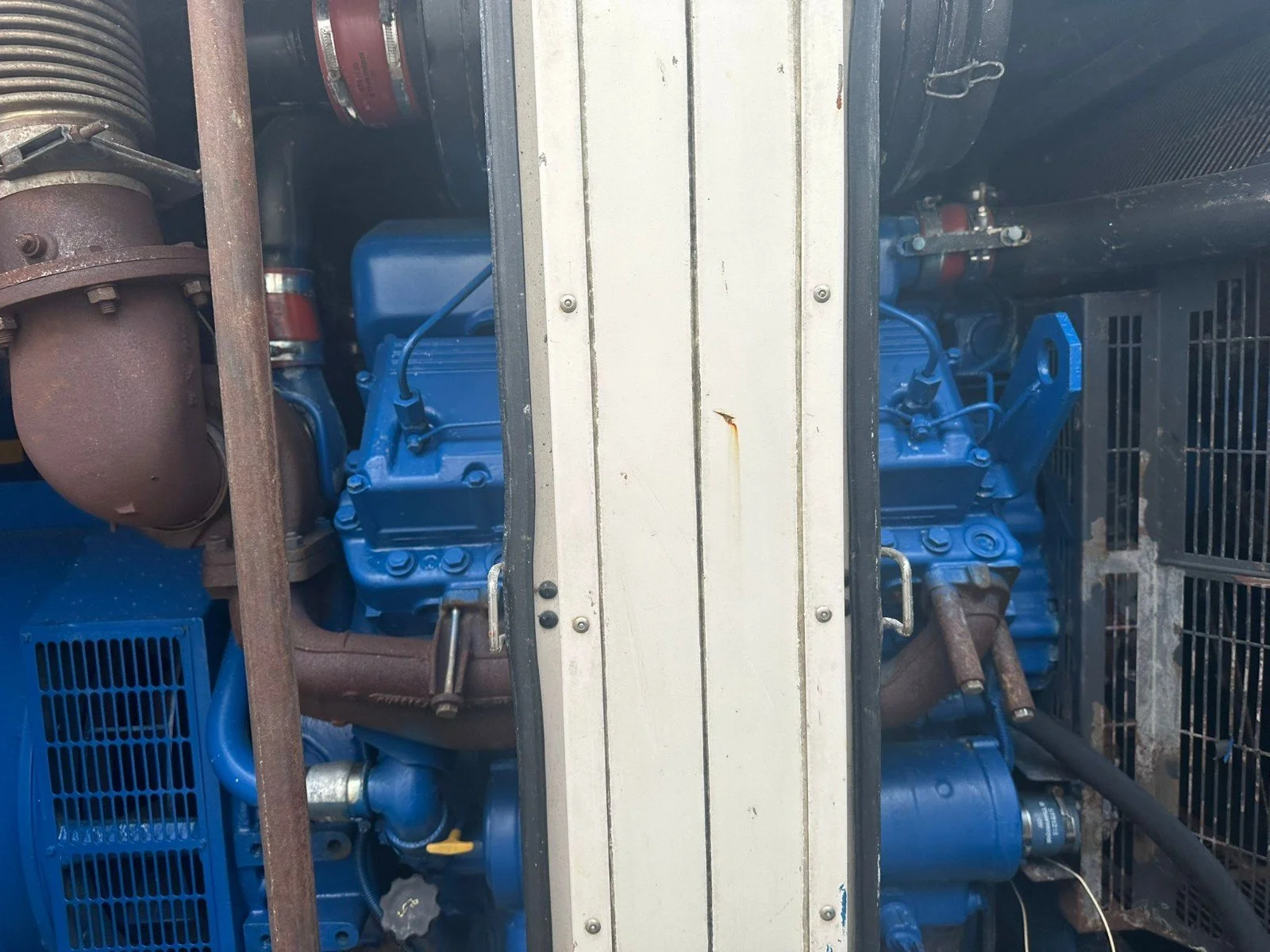 A close-up view of two blue industrial engines or generators inside a metal enclosure, with various pipes and components visible.