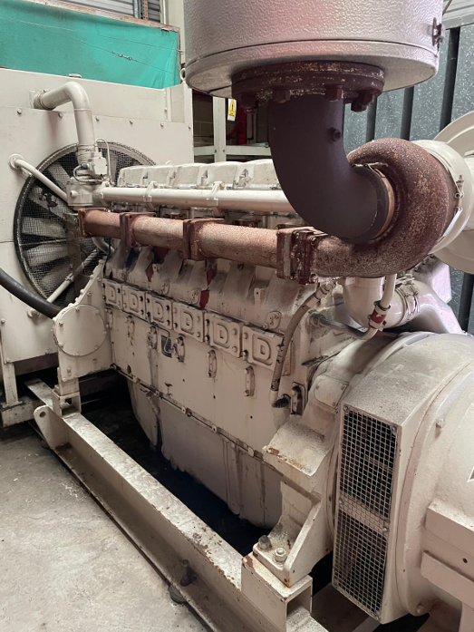 A large industrial generator with a rusted exhaust pipe on top, situated indoors on a concrete floor.