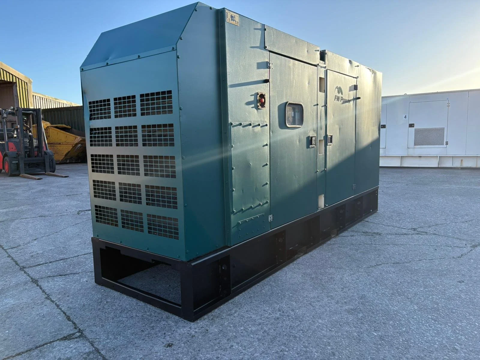 Large industrial generator painted green on concrete surface outdoors with warehouse and forklift in background during daylight.