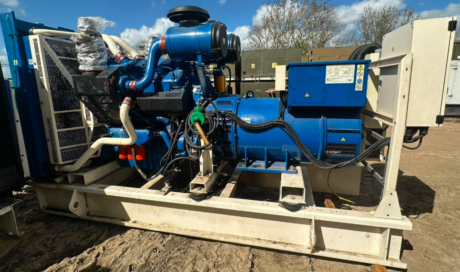 Industrial generator or engine with blue and black components, white pipes, and warning labels, installed outdoors on a metal frame.