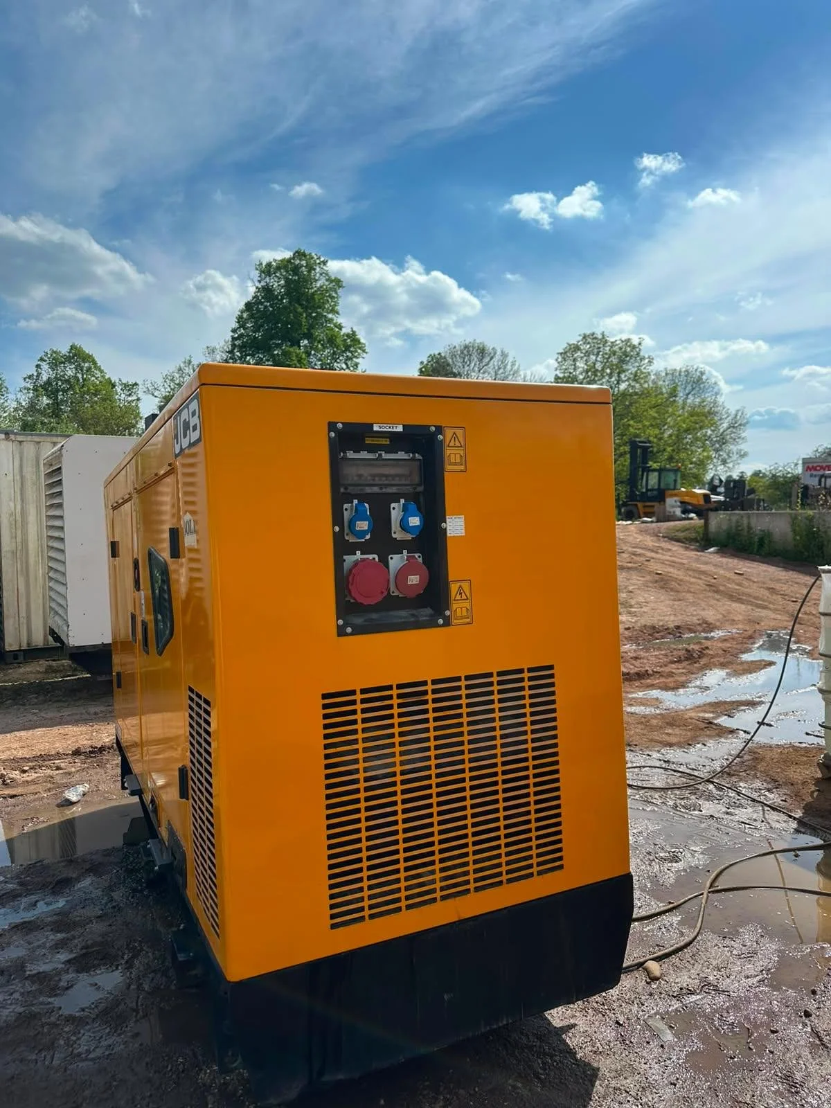 A bright yellow portable generator with front panel displaying electrical outlets, set on a construction site with muddy ground, trees, and construction equipment in the background under a partly cloudy blue sky.