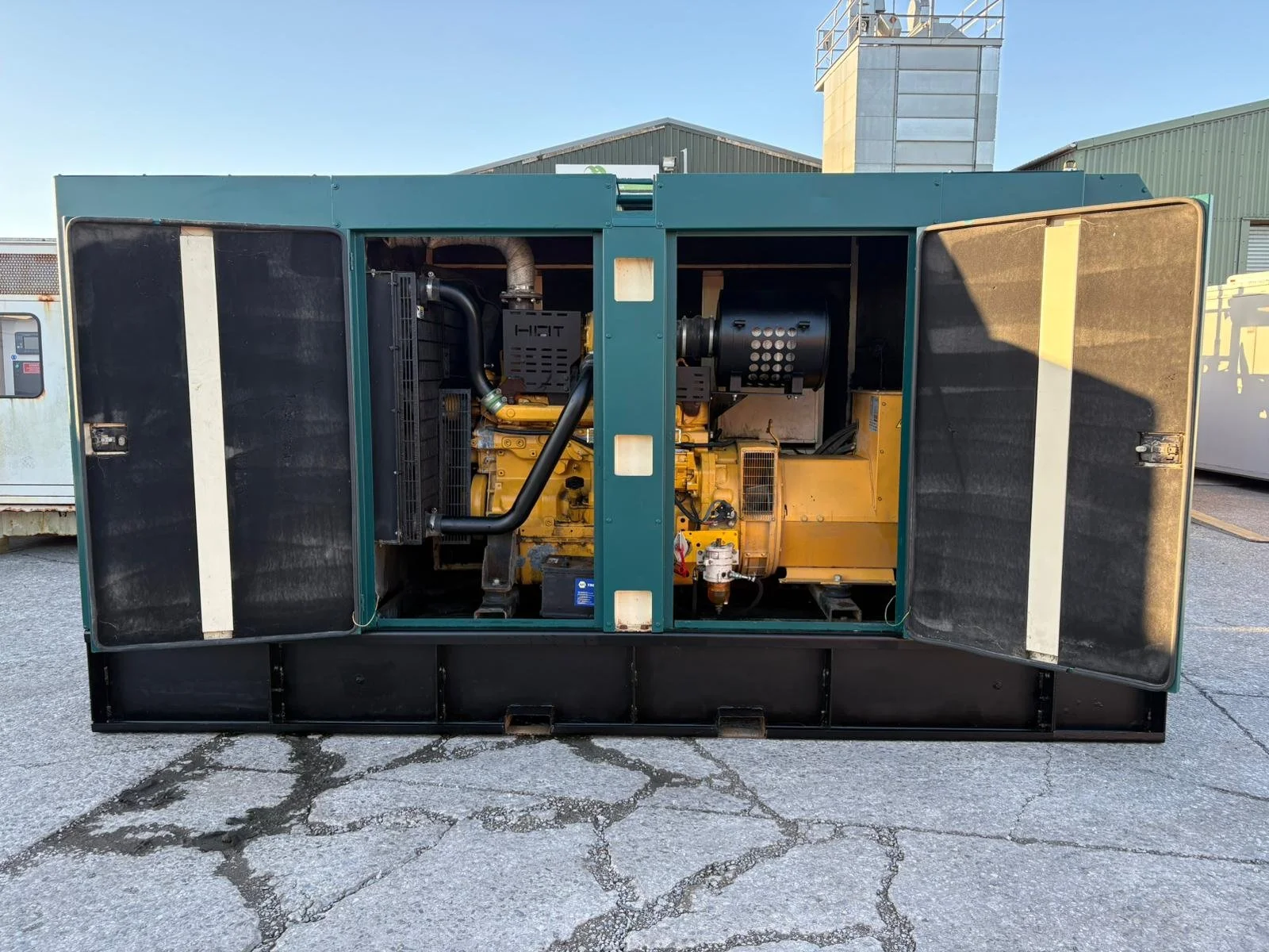 Open generator with yellow engine and black exhaust unit, metal shelter, vents, and control components, outdoors on cracked concrete surface.