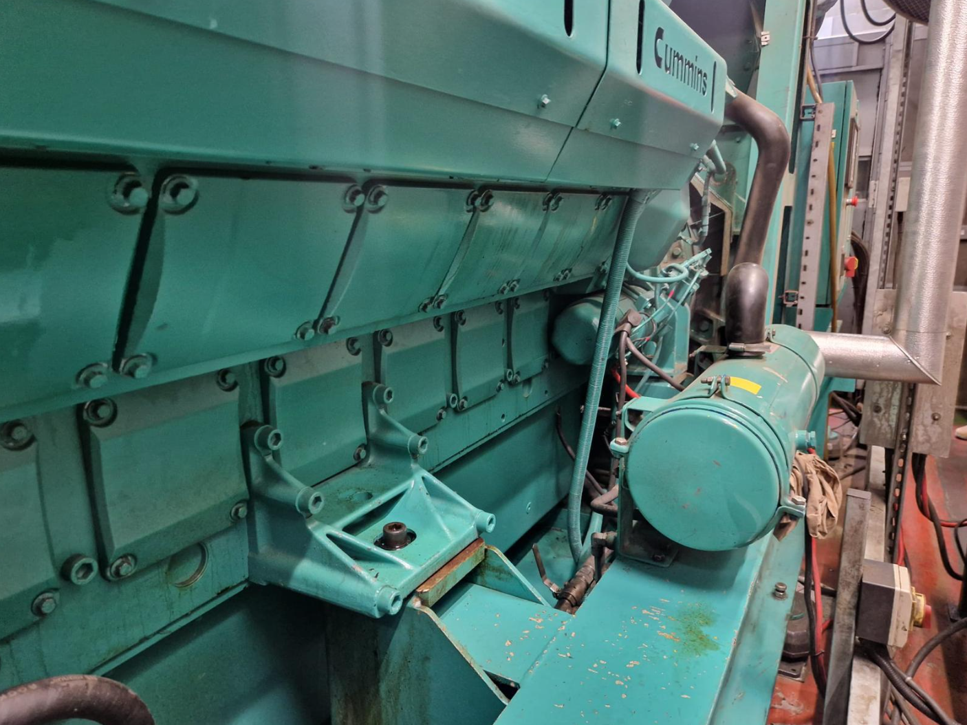 Large industrial turquoise-colored engine or generator with visible bolts, pipes, and electrical wiring, situated in an industrial setting.