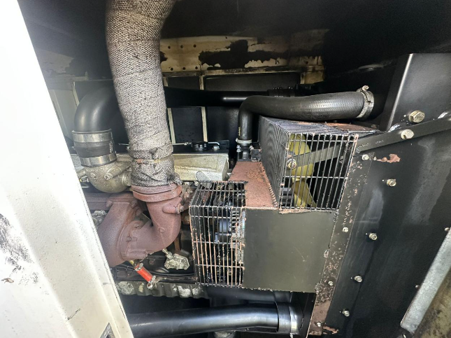 Close-up of machinery with metal pipes, a heat shield wrapped with insulation, a fan, and protective metal grilles.
