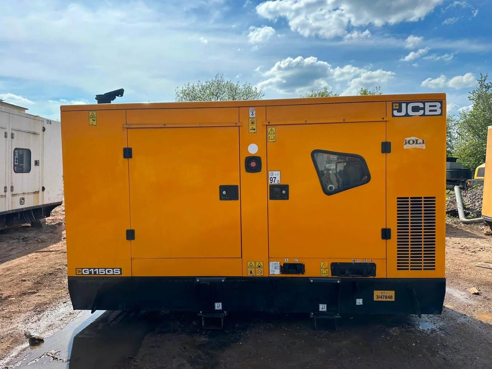 A large yellow industrial generator with black base and controls, situated outdoors on dirt ground, under a blue sky with some clouds.