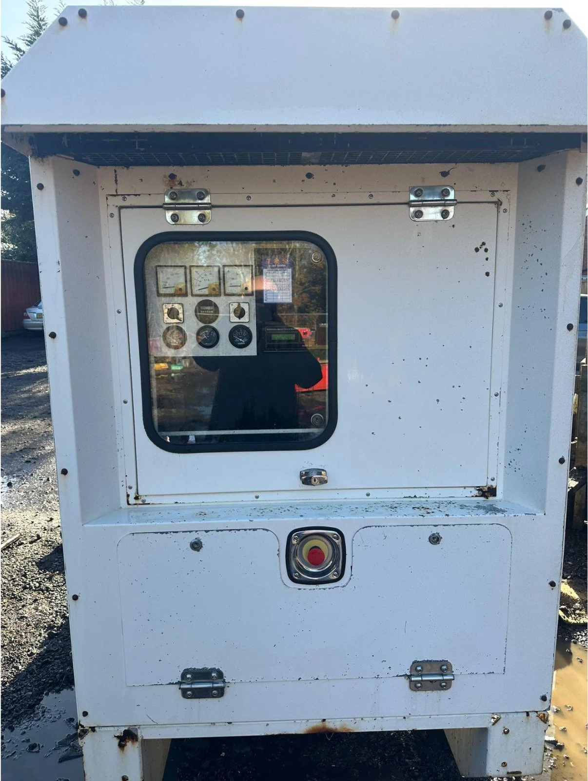 A white electrical or utility box with a window and buttons on the front, showing gauges and controls inside, set outdoors on a muddy ground.