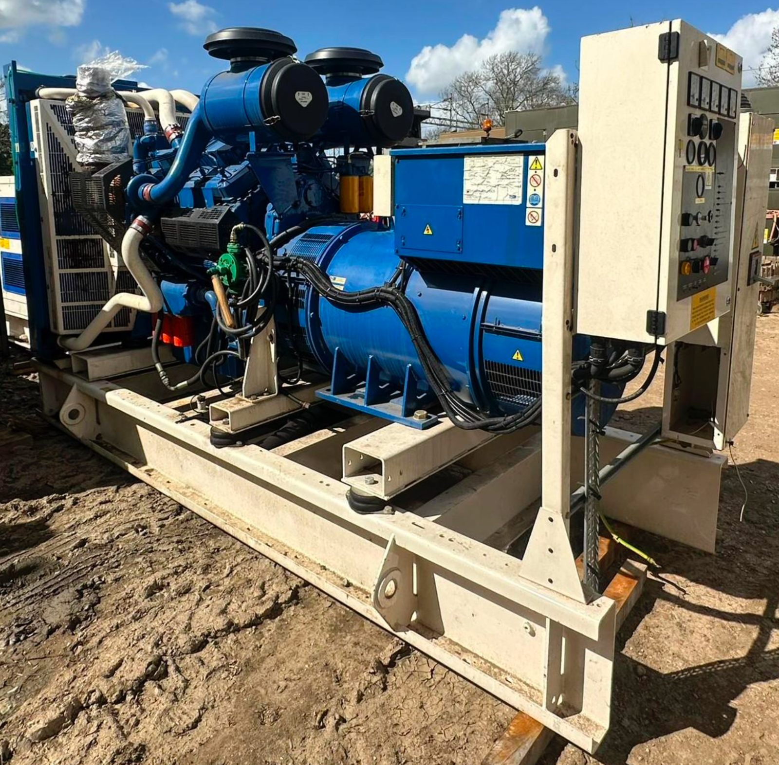 Industrial mechanical equipment with blue and black components mounted on a white metal frame, outdoors on dirt ground with trees and cloudy sky in background.