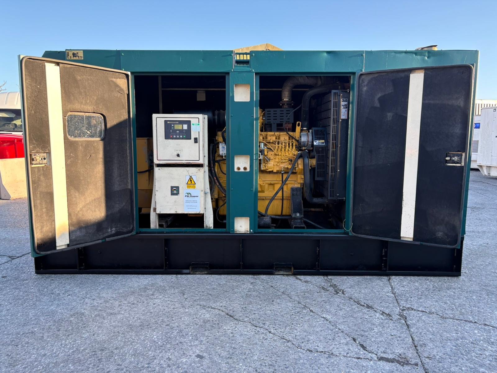 Open generator enclosure showing generator and control panel inside a large metal box, outdoors on concrete surface.