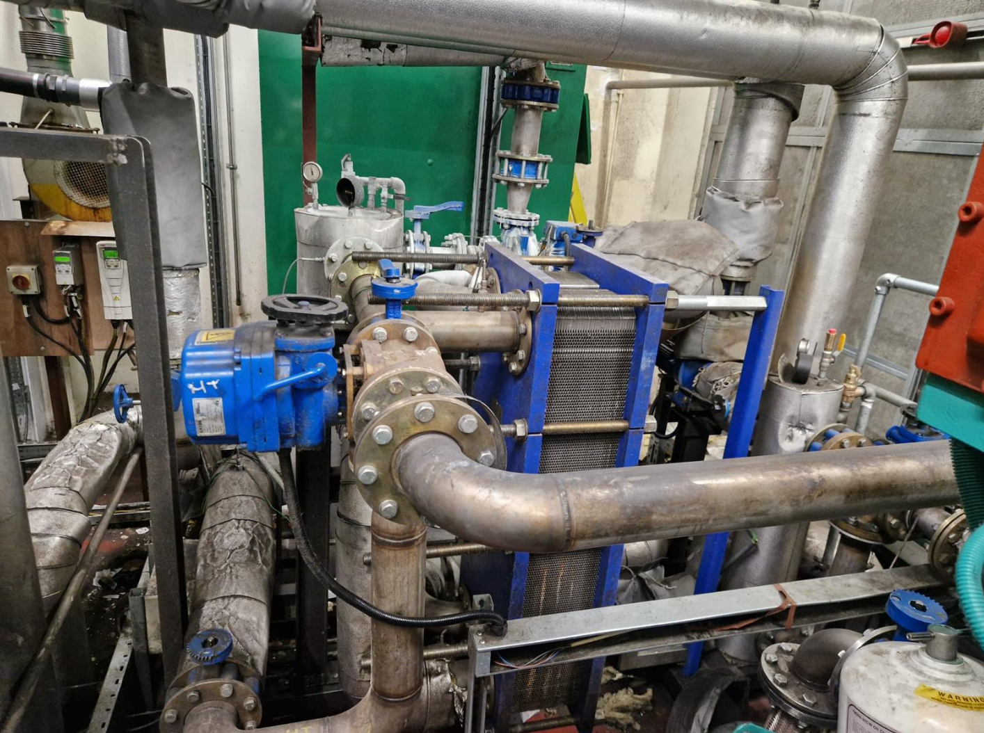 Industrial machinery with large pipes, gauges, and a blue pump, part of a mechanical or HVAC system in a utility room.