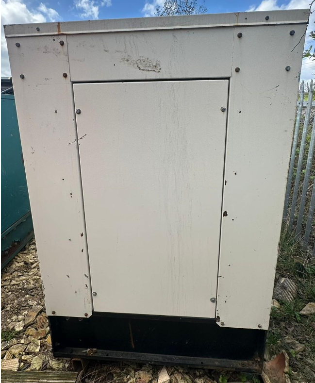 An outdoor electrical or utility box with a closed door, located on rough ground with debris, and surrounded by a metal fence.