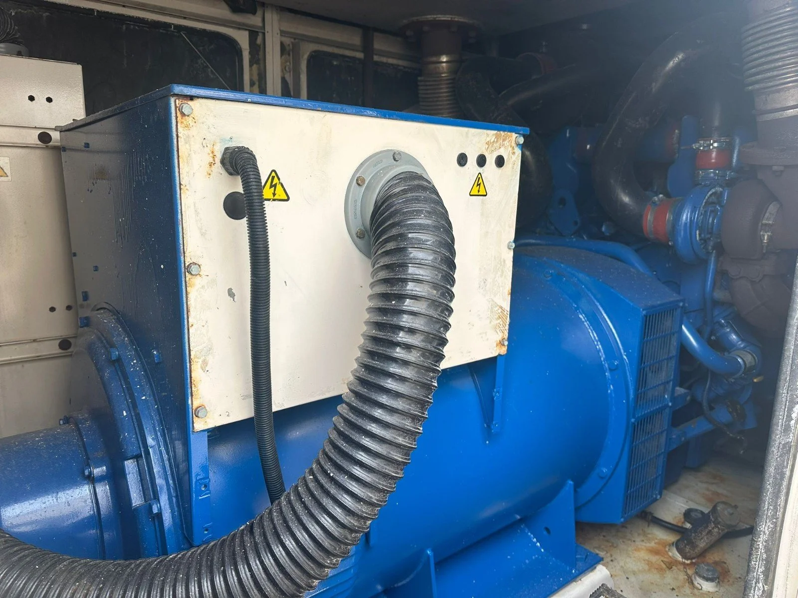 Close-up of industrial machinery featuring a large black flexible hose connected to a metal panel with yellow warning symbols, part of a blue engine or generator with various pipes and components.